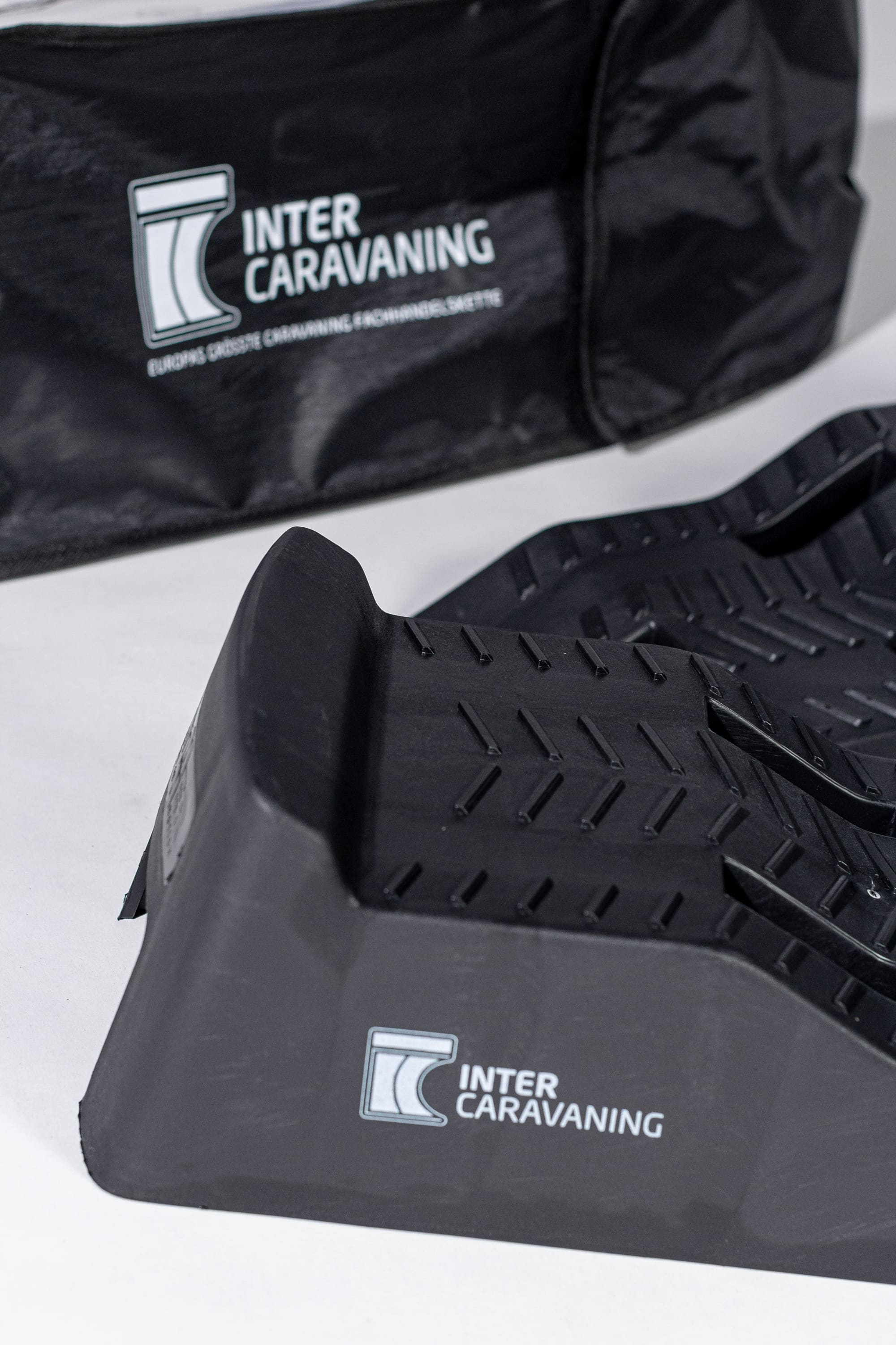 Schwarze Auffahrkeile Equalizer XL InterCaravaning Edition mit rutschfester Oberfläche; im Hintergrund Tragetasche mit InterCaravaning-Logo