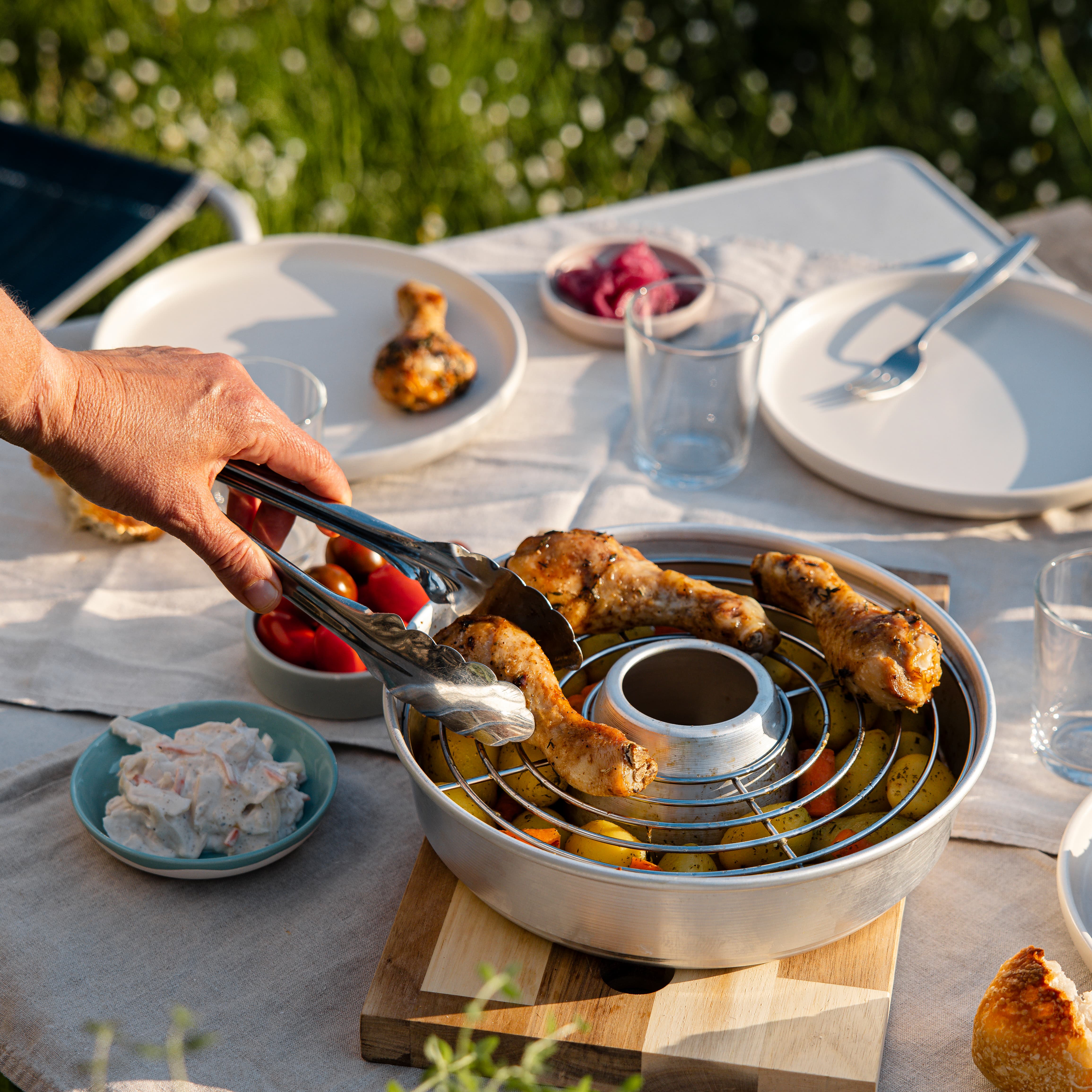 Freilufttisch mit gegrillten Hähnchenteilen in einem Metall-Dampfkorb, darunter Kartoffeln, daneben Tomaten in einer Schale, Glas Wasser, Teller und Besteck auf heller Tischdecke