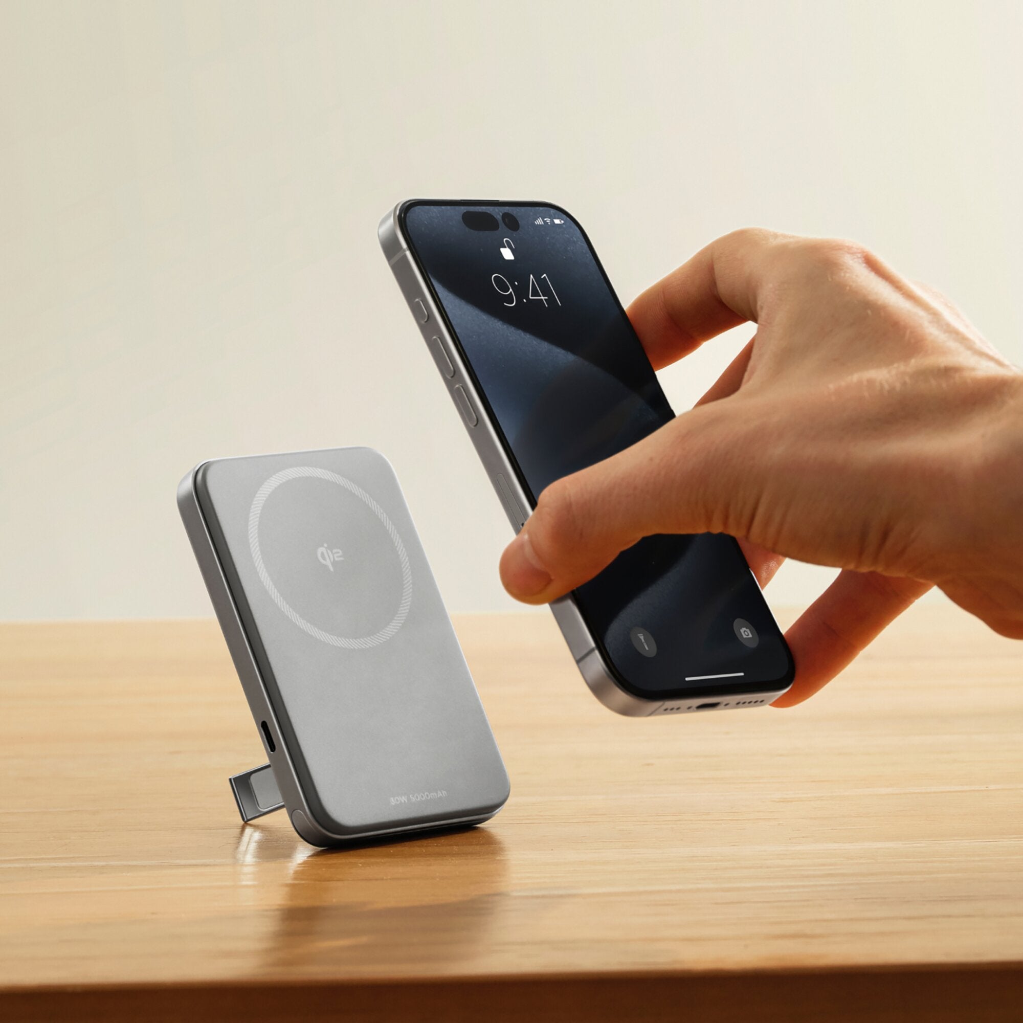 Hand hält ein Smartphone nahe an einer magnetischen Powerbank Rapid, 5000 mAh, daneben liegt eine graue Qi-Ladeplatte auf einem Holzuntergrund
