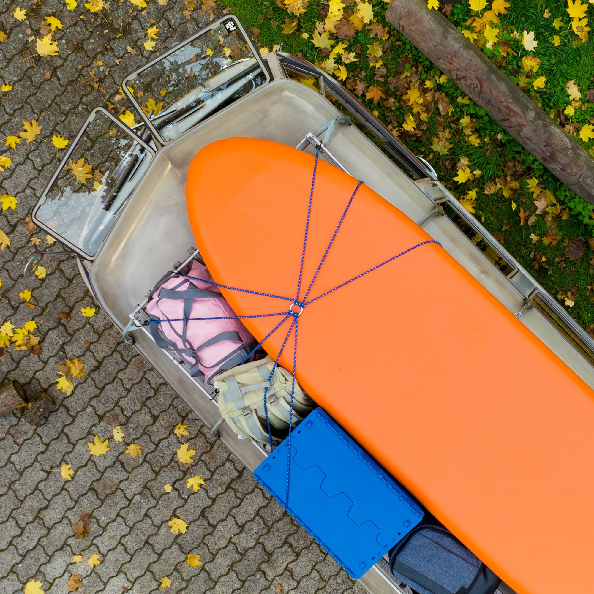 Vogelperspektive auf eine geöffnete Autoladefläche; ein orangefarbener Gegenstand ist durch eine blaue Gepäckspinne gesichert, Herbstlaub darum