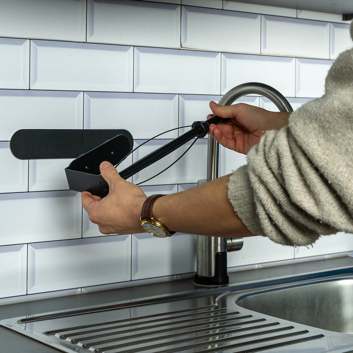 Nahaufnahme eines schwarzen Magnet-Küchenrollenhalters mit Metall-Leiste, den eine Hand festhält, Hintergrund Spüle und weiße Fliesen