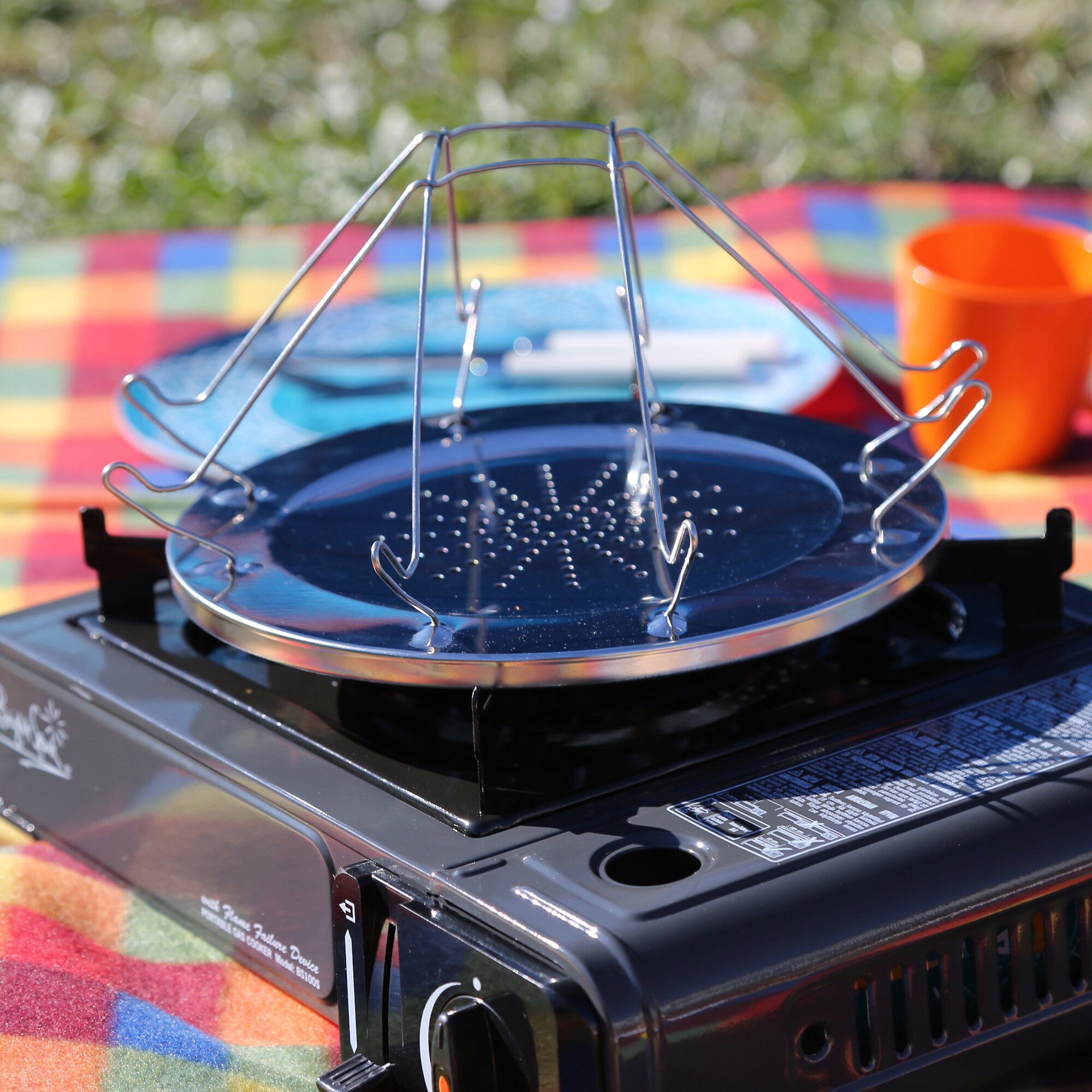 Nahaufnahme von Edelstahl-Campingtoaster mit Drahtkorb über einer Grillplatte, draußen auf bunter Picknickdecke