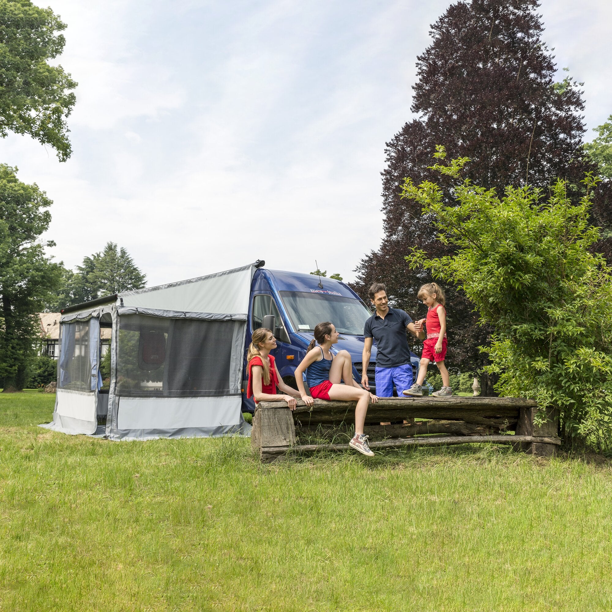 Blauer Transporter mit seitlichem grauen Markisenzelt Privacy Room F80, Seitenverkleidung mit Netzfenstern, fünf Personen in Freizeitkleidung stehen und sitzen davor auf einer Holzbank im Garten