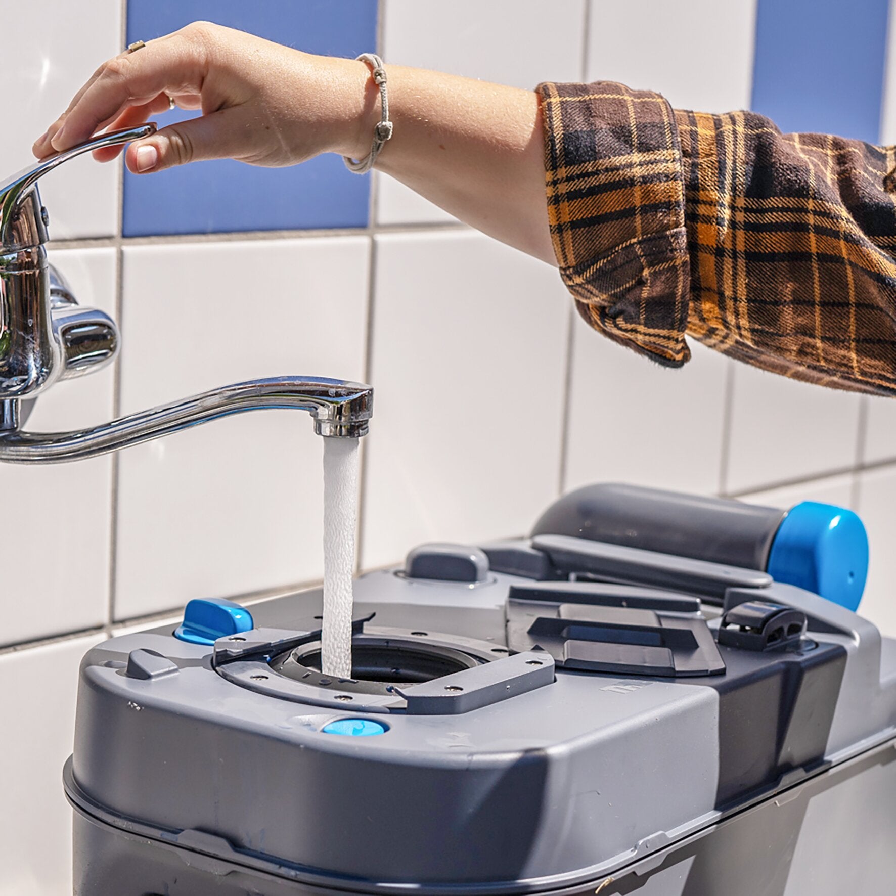 Eine Person mit kariertem Hemdärmel betätigt einen Wasserhahn, Wasser fließt in einen grauen Behälter mit blauem Deckel, vor Fliesen