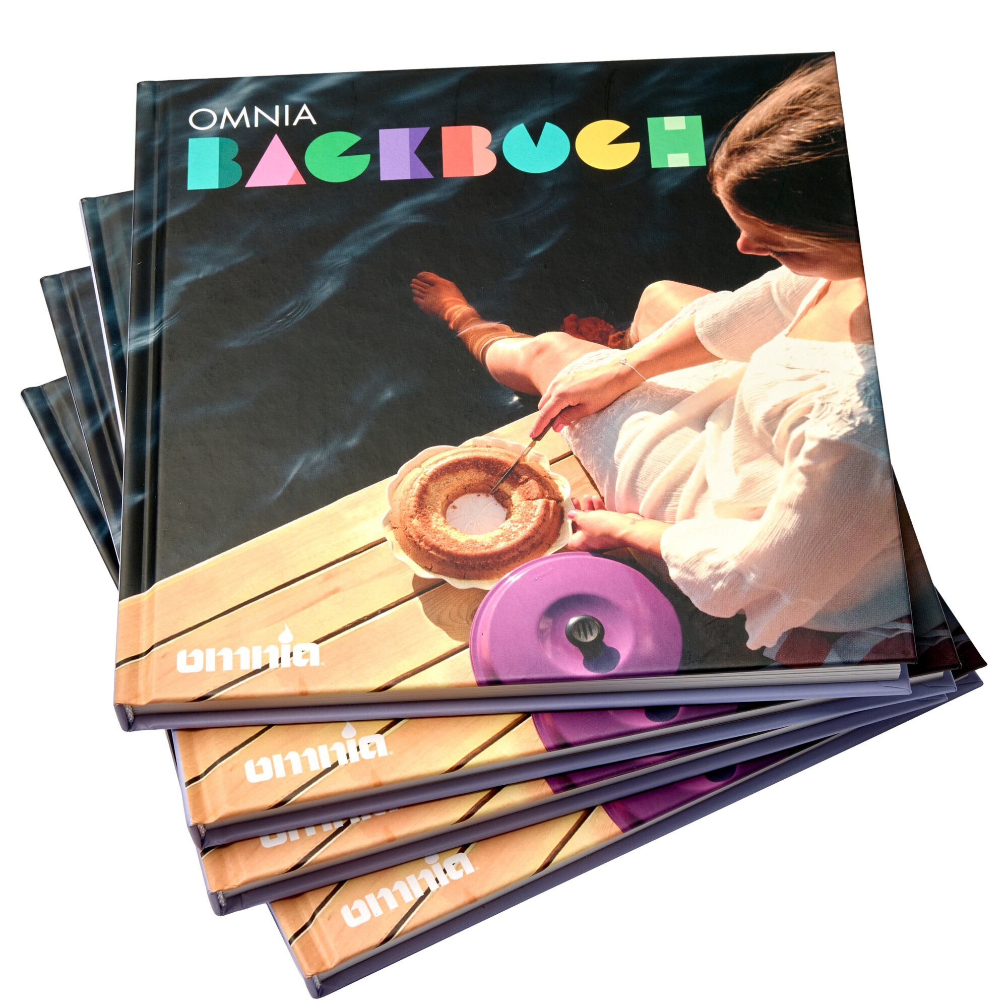 Mehrere Exemplare des Omnia Backbuchs, bunte Buchstaben auf dem Cover, neben einer Person beim Backen und eine runde Backform auf Holztisch
