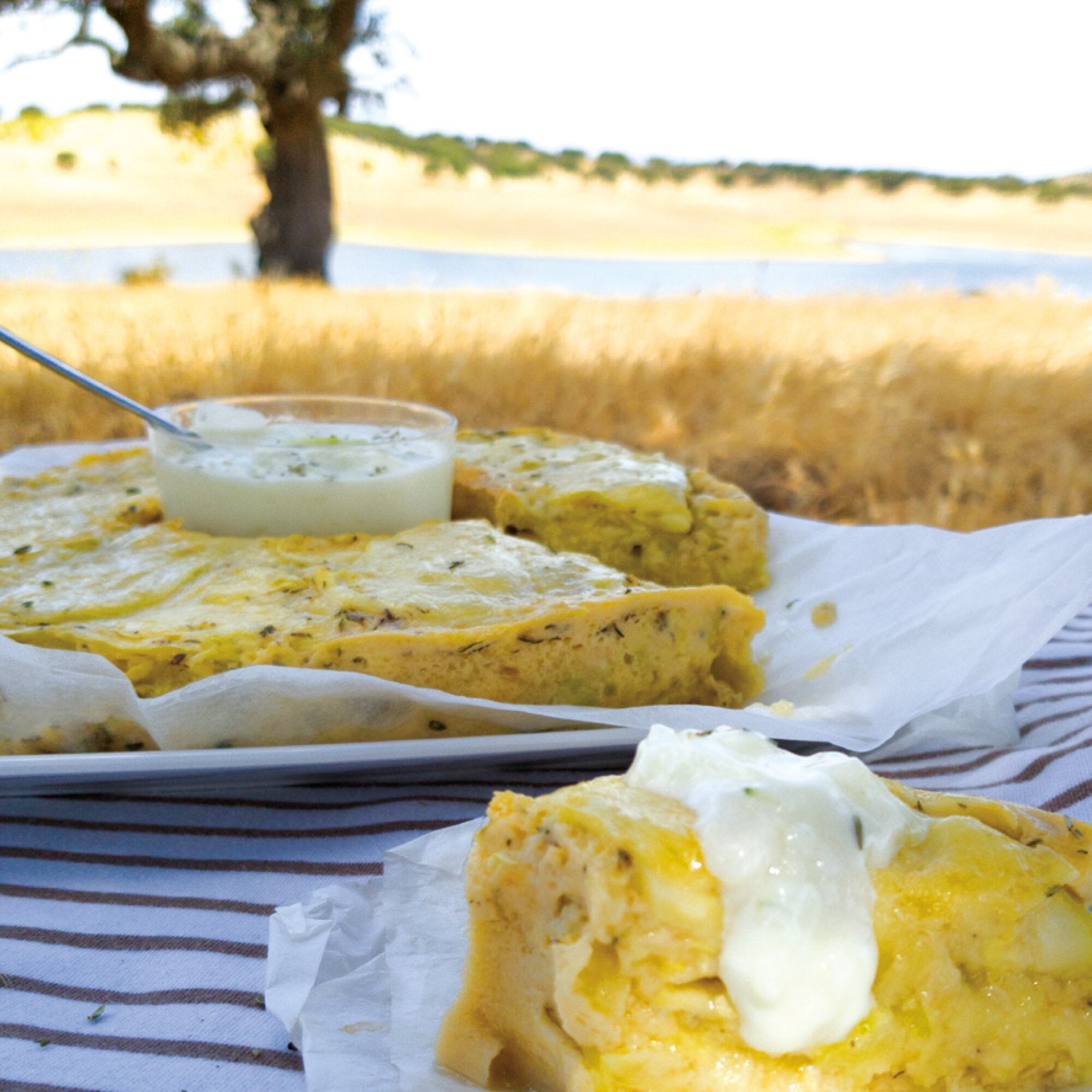 Quiche-Scheiben auf einer weißen Platte mit Backpapier, daneben ein Glas mit cremigem Dip; im Hintergrund eine sonnige Landschaft mit Baum und Fluss
