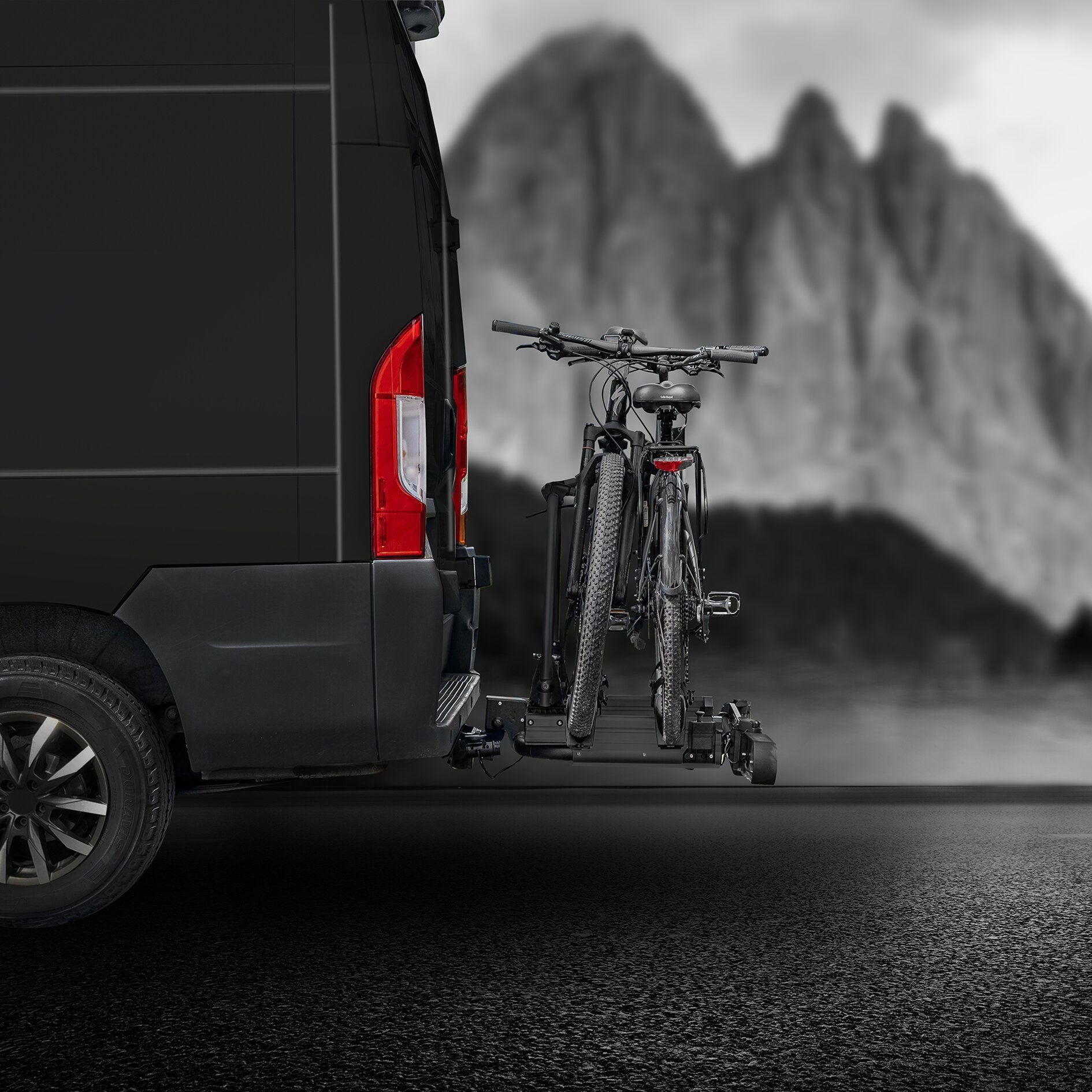 Schwarzer Van mit montiertem schwarzen Fahrradträger an der Anhängerkupplung, zwei Mountainbikes hängen am Heckträger, Landschaft im Hintergrund