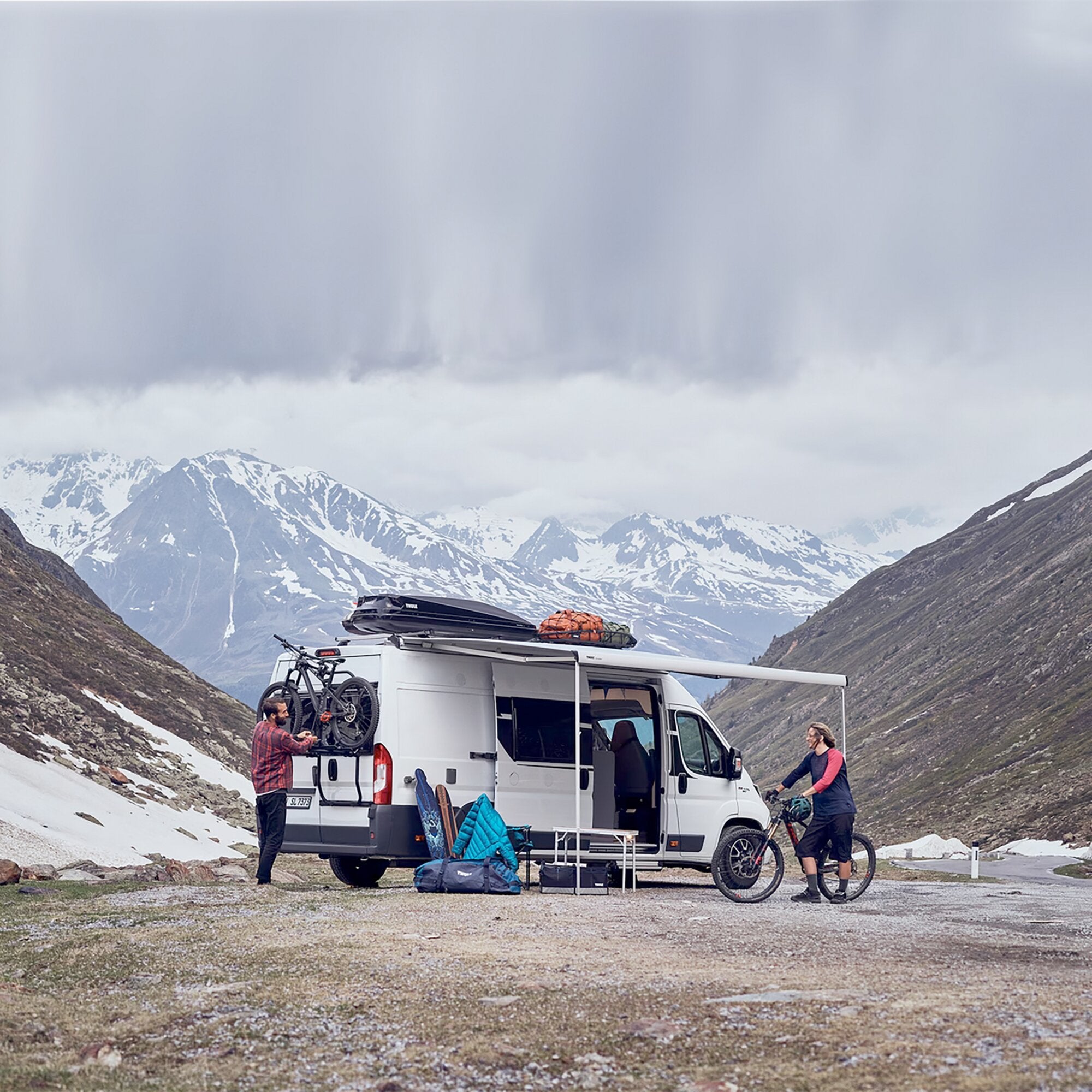 Weißer Camper-Van mit schwarzem Thule Elite Van XT Fahrradträger auf dem Dach, bergige Schneelandschaft im Hintergrund, zwei Personen daneben