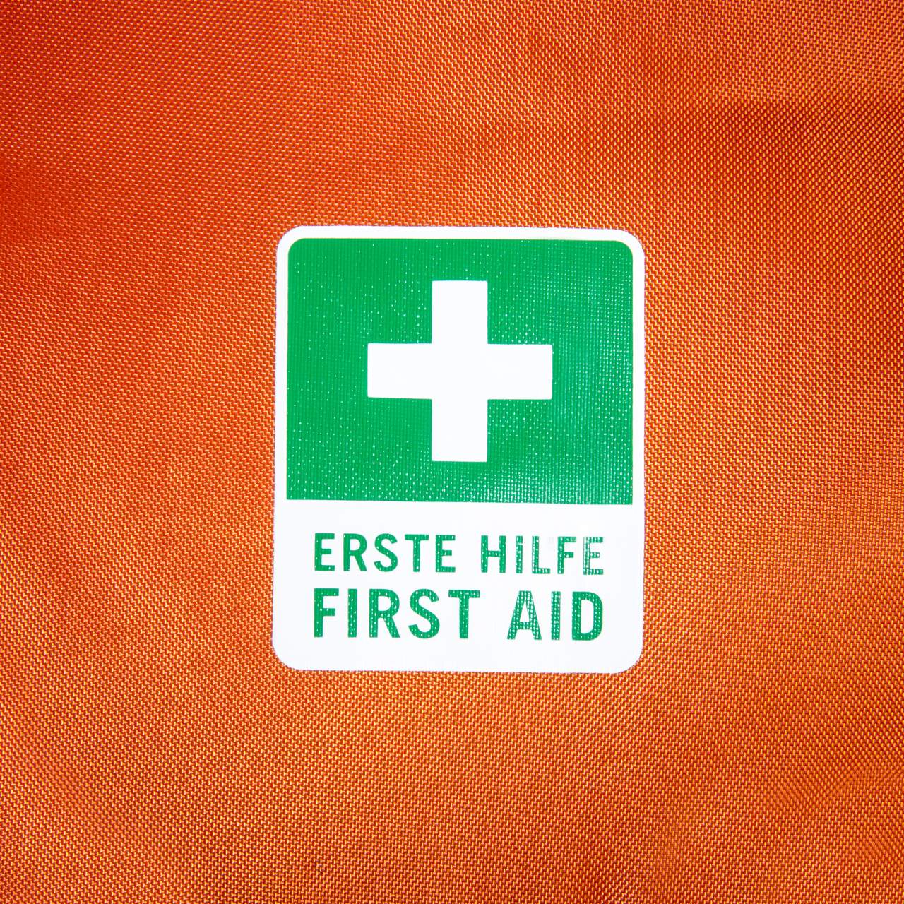 Orangefarbener Stoff mit aufgenähtem grün-weißem Erste-Hilfe-Schild und Kreuzsymbol.
