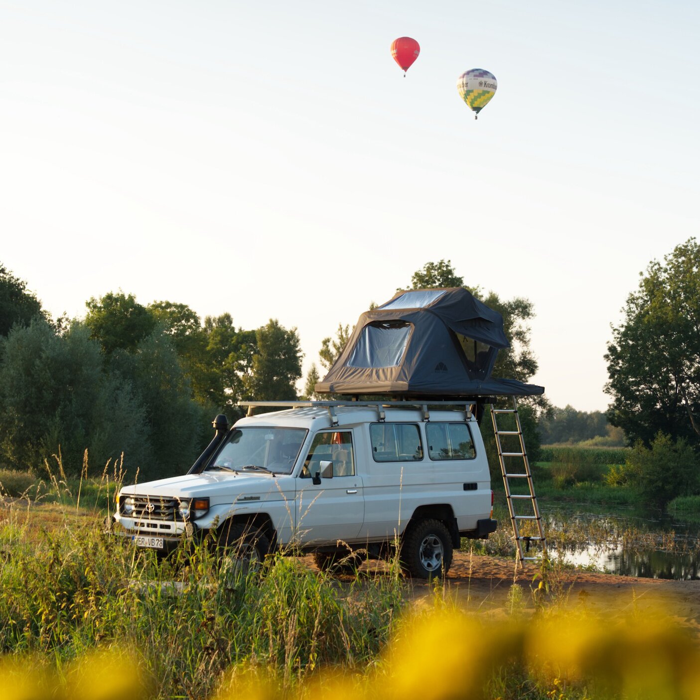 Weißer Geländewagen mit aufgebautem Dachzelt Roof Space 2 auf dem Dach, Leiter seitlich, Uferlandschaft und zwei Heißluftballons am Himmel