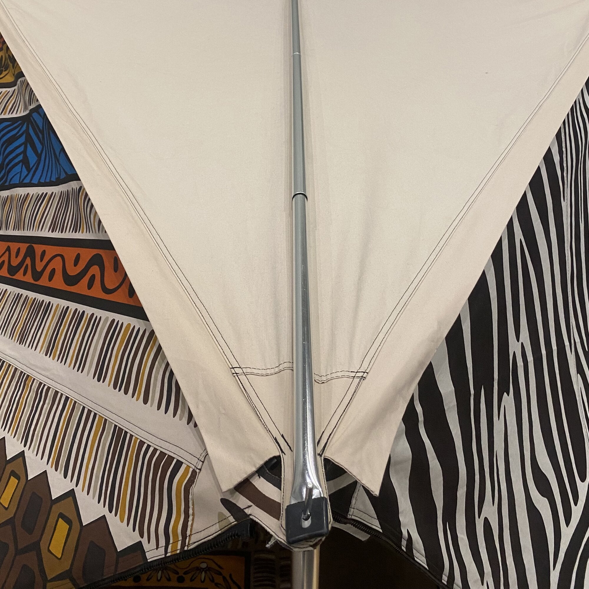 Mittig stehender silberner Aluminium-Spannstab durch beige Stoffplane; sichtbare Nähte, links farbige Muster, rechts zebraartige Streifen