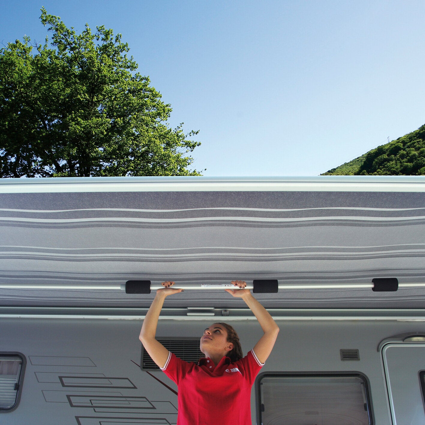 Person in rotem Shirt hebt eine silberne Querstange Magicrafter 400 unter der Caravan-Decke eines Wohnmobils, draußen blauer Himmel, Bäume und Hügel