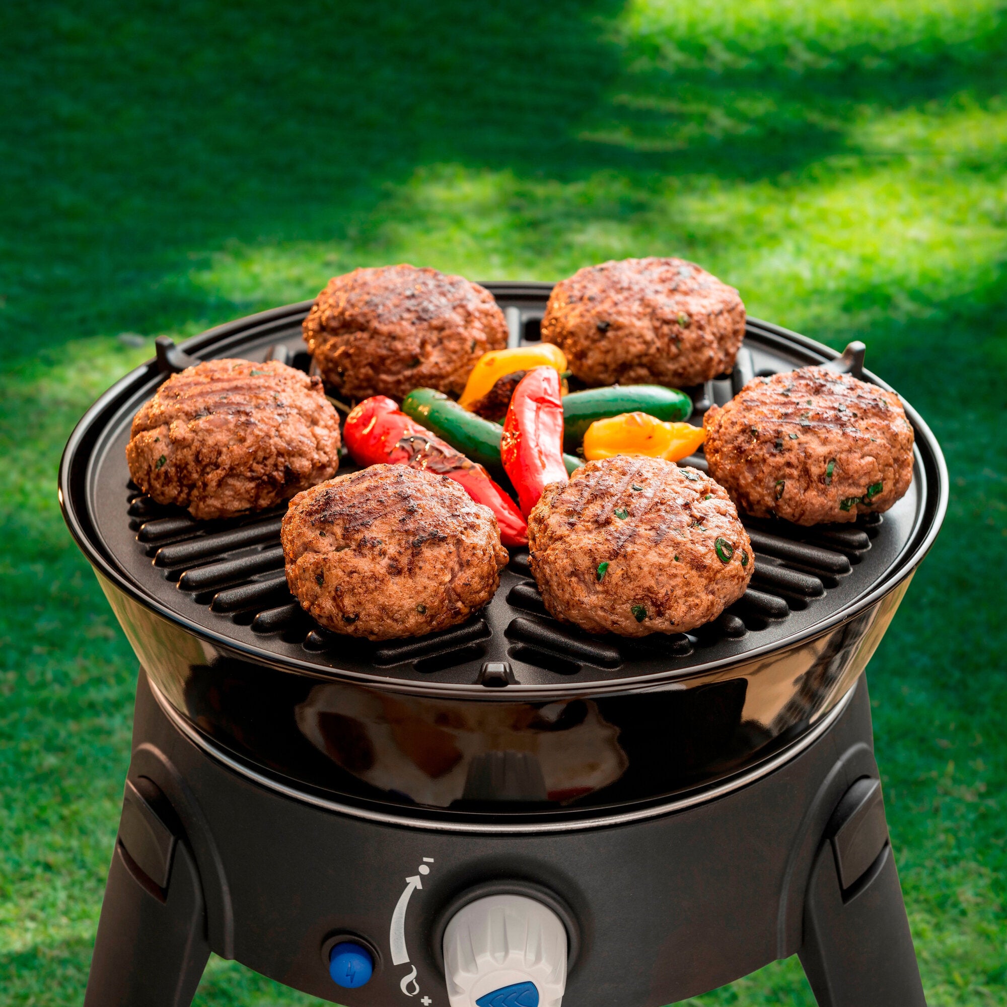 Runder Gasgrill mit offenem Deckel, darauf gegrillte Hamburger-Patties und bunte Paprika, Hintergrund grüne Wiese