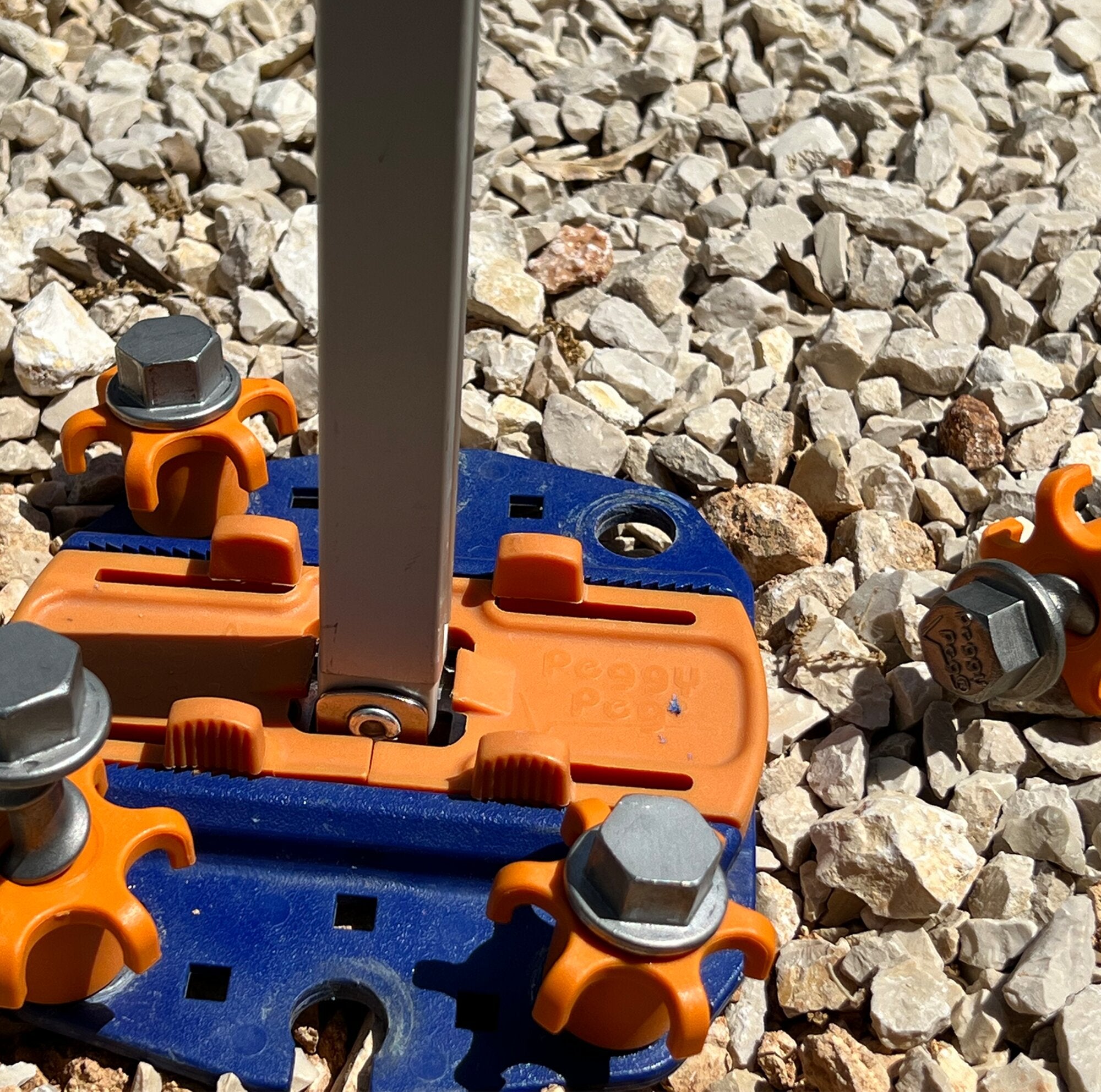 Detailaufnahme des PP Schraubhering Peg&Stop HeavyDuty: orange Kunststoff-Clips, grauer Metallpfosten, blaue Grundplatte auf Kiesboden