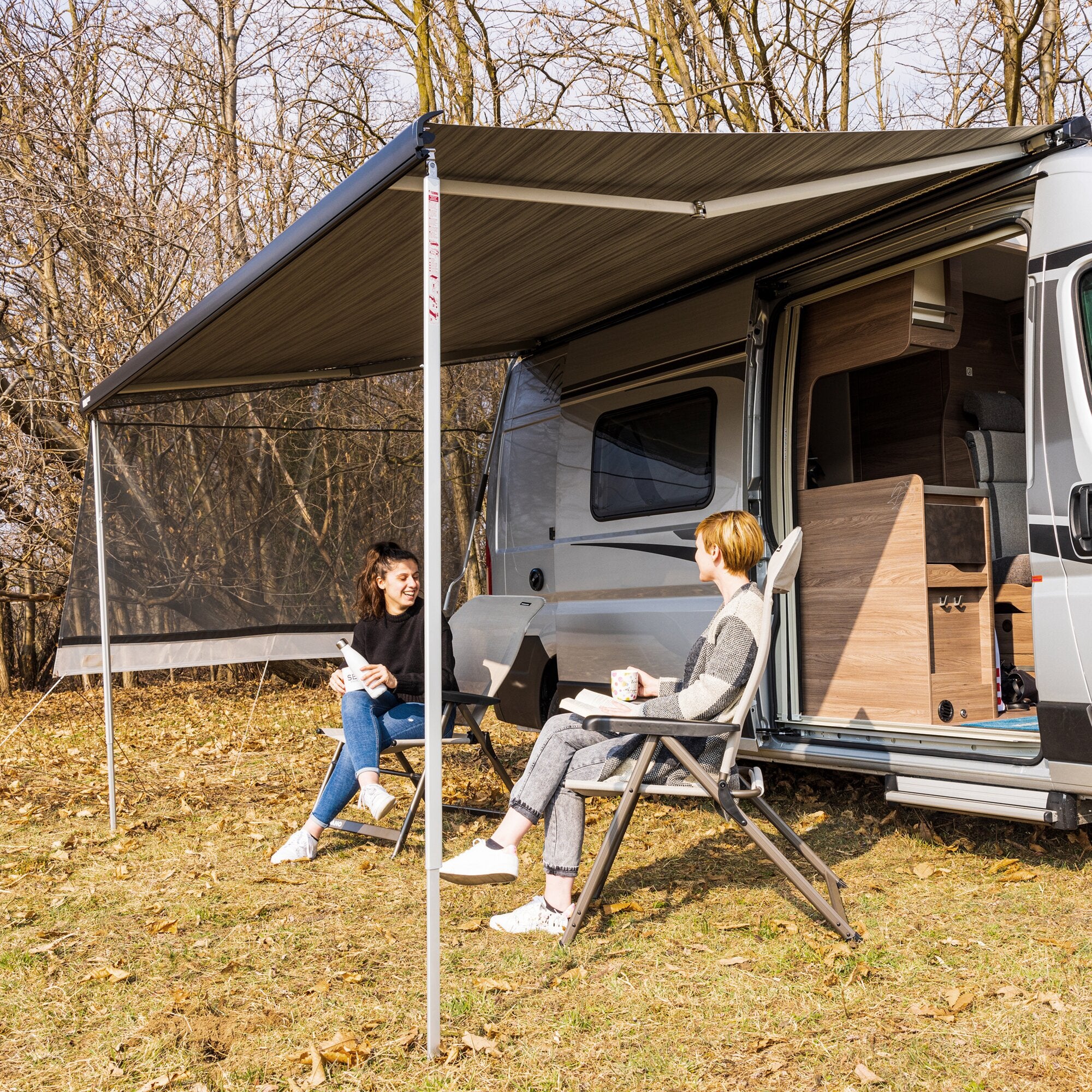 Ausgefahrene Sun-View-Seitenwand am Wohnmobil mit Markise, Stützbeine mit Maßskala, zwei Personen sitzen in Klappstühlen vor dem Innenraum