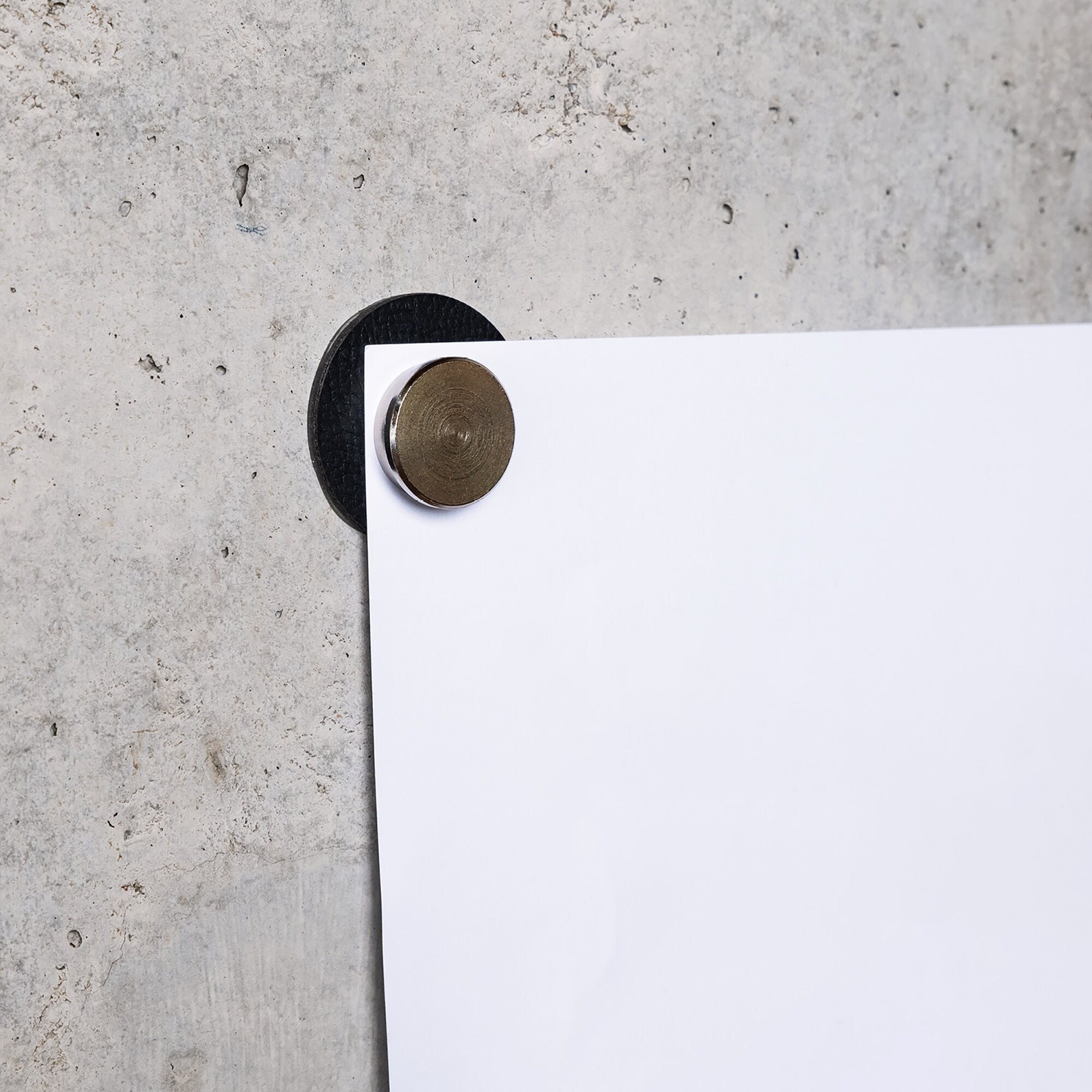 Runder bronzefarbener Magnetkern auf einer schwarzen Unterlage hält ein weißes Blatt Papier an eine raue Betonoberfläche