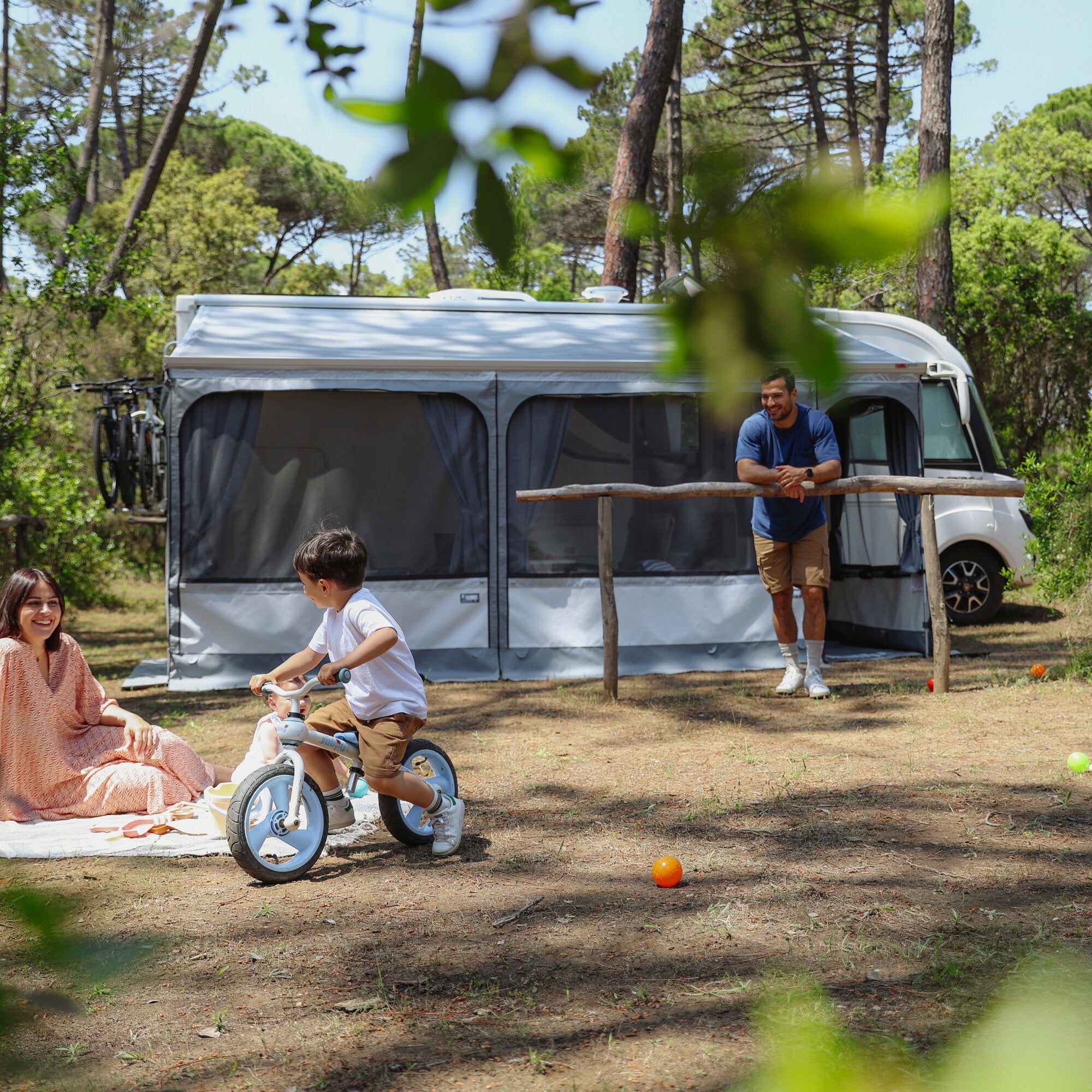 Großes Wohnmobil mit grauem Markisenzelt Privacy Room F45, 300 Large; Mann neben der Tür lächelt, Kind auf Dreirad im Vordergrund, Bäume im Hintergrund
