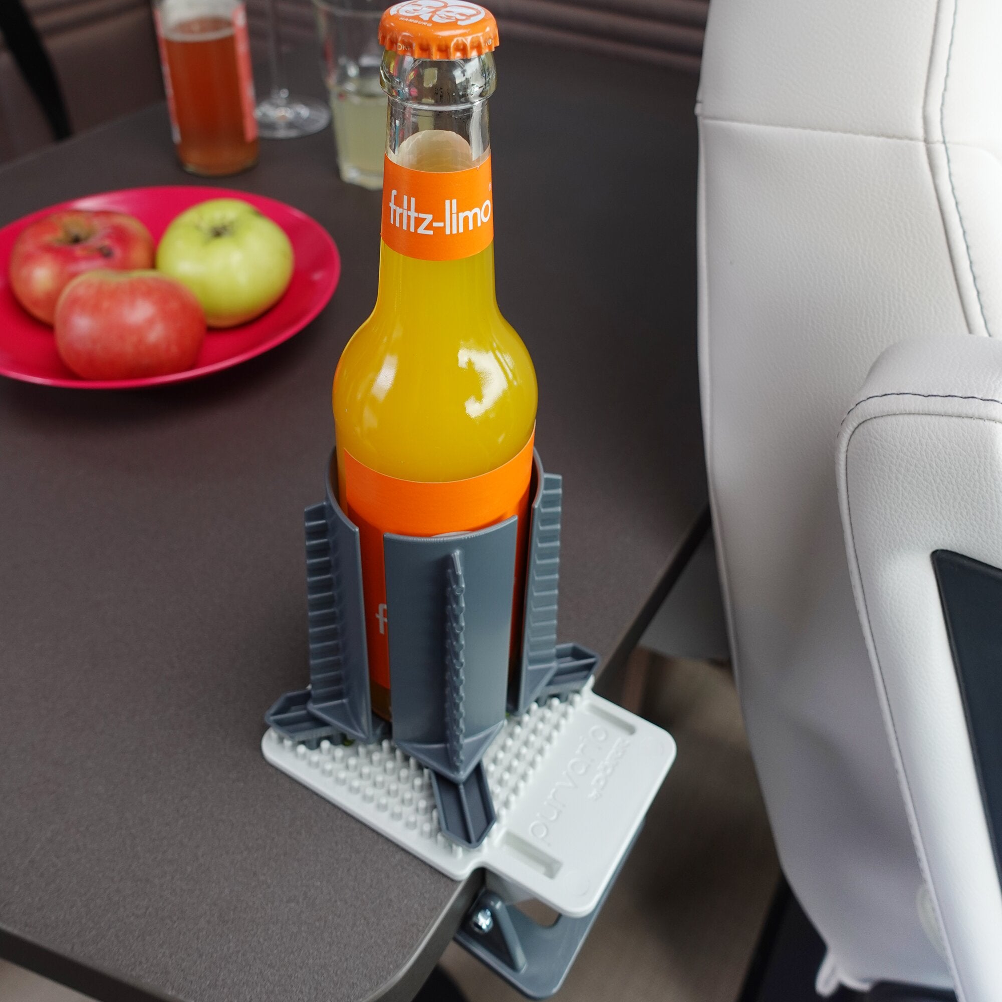 Clip Clap Getränkehalter hält Fritz-Limo Flasche senkrecht, grauer Klemmhalter mit orange Bändern am Tischrand; Obstplatte mit Äpfeln