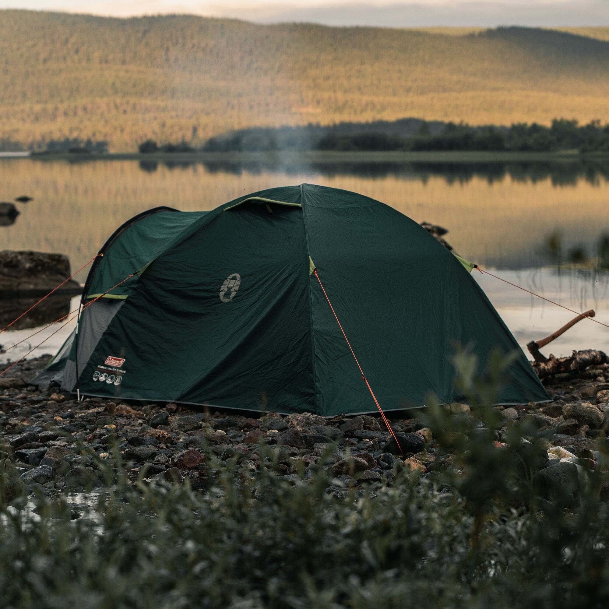 Grünes Kuppelzelt Kobuk Valley Plus, 3-Person, am Ufer eines kiesigen Sees; orange Abspannseile, ruhiges Wasser und sanfte Hügel im Hintergrund