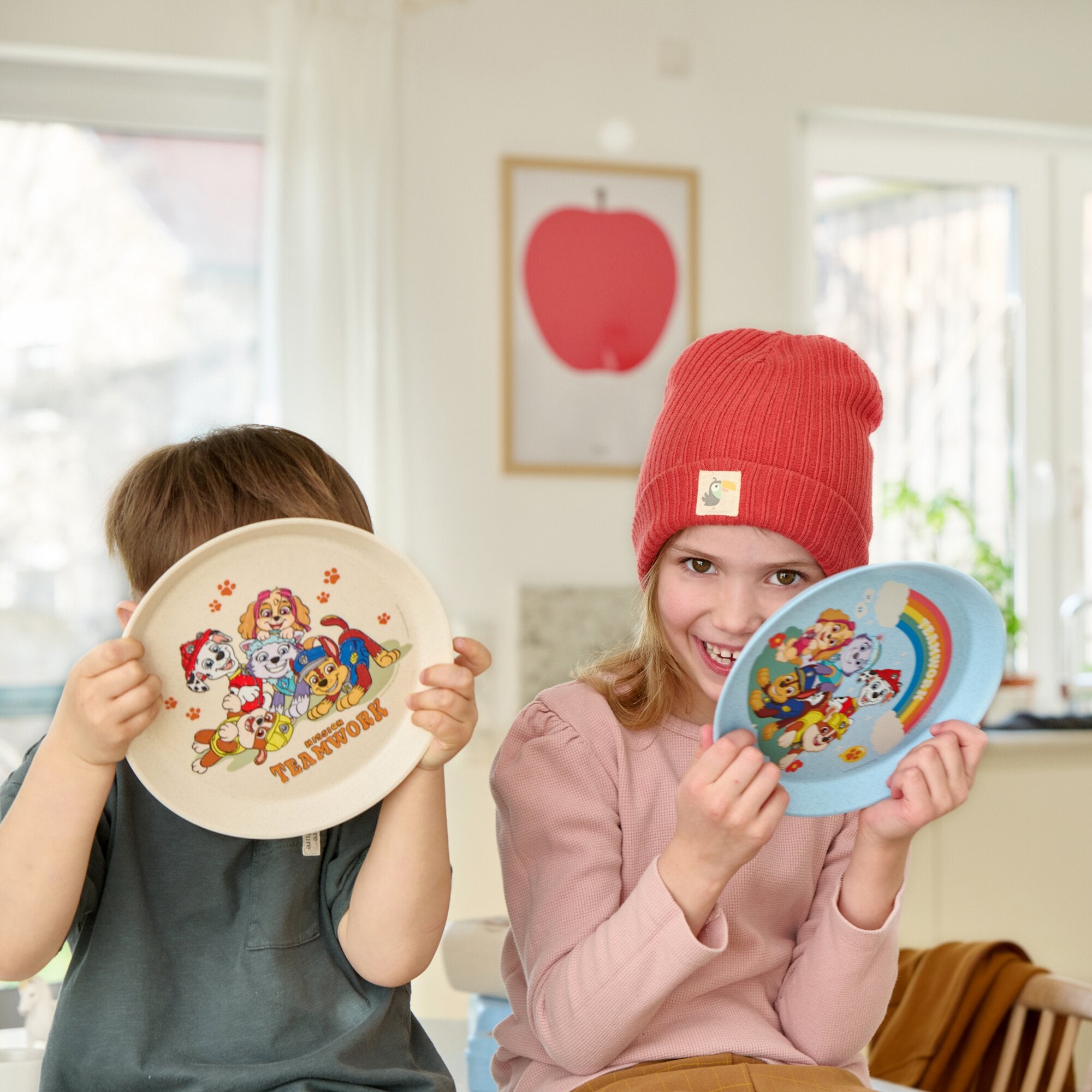 Zwei Kinder zeigen Paw Patrol Kindergeschirr: links weißer Teller mit bunten Charakteren, rechts blauer Teller mit Regenbogenmotiven