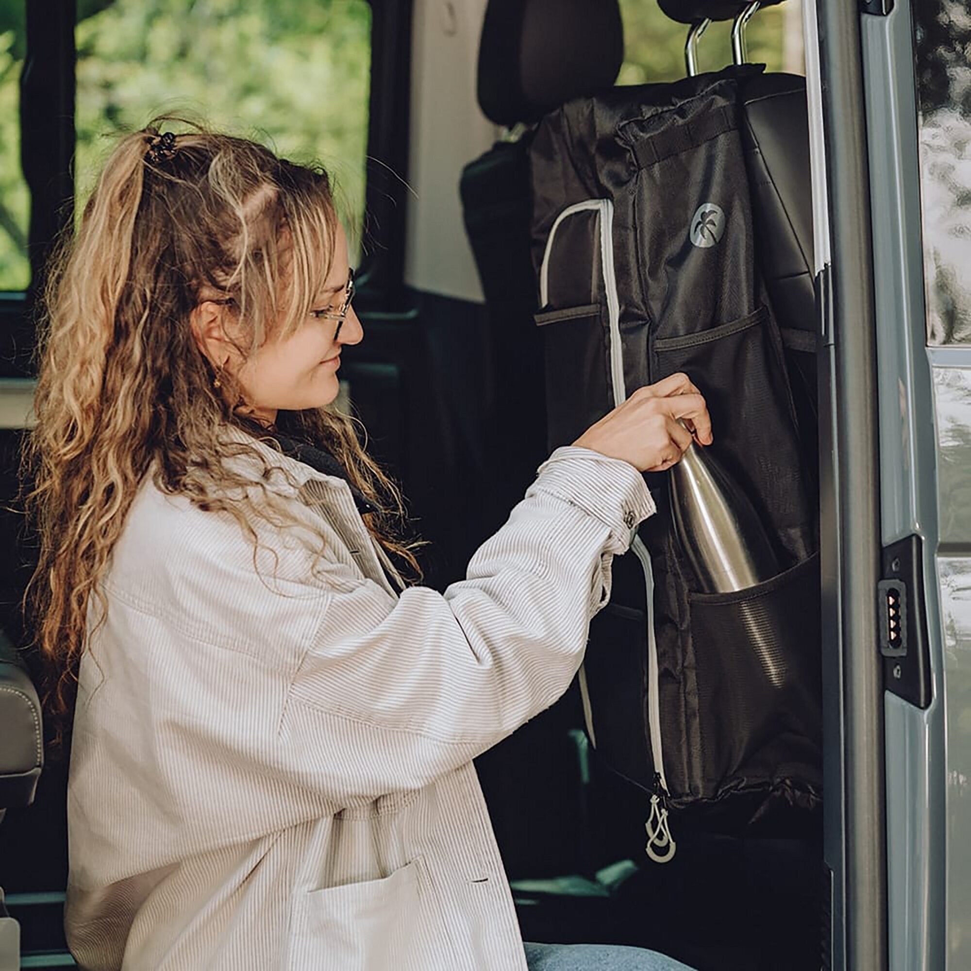 Person mit lockigem Haar sitzt in einem VW-Van und befestigt eine schwarze seatBOXX Organizer-Tasche an der Rückenlehne des Vordersitzes, hält eine Edelstahlflasche