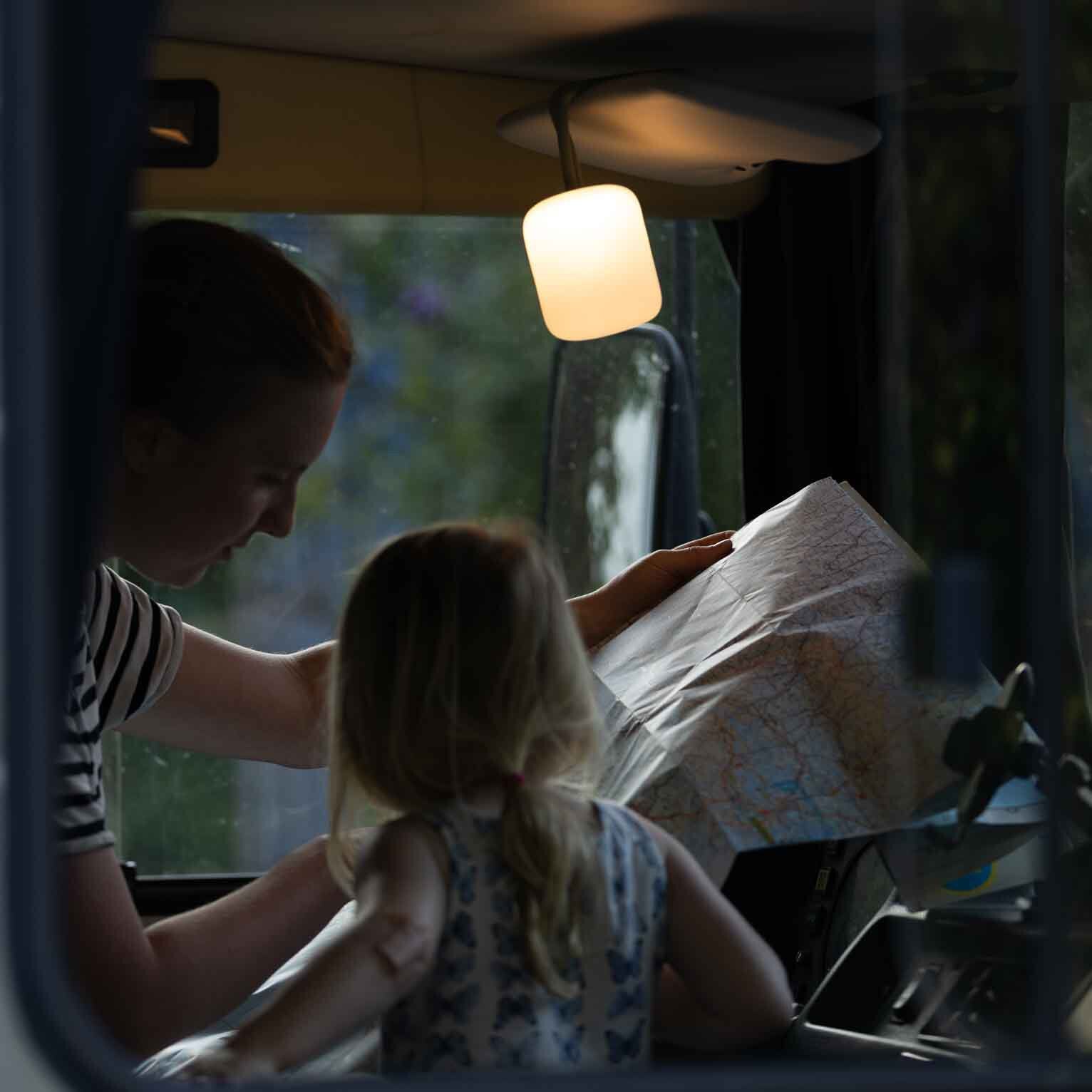 Campinglaterne Floppy aus Silikon in Orange hängt von der Fahrzeugdecke und spendet warmes Licht, während Personen eine Karte betrachten