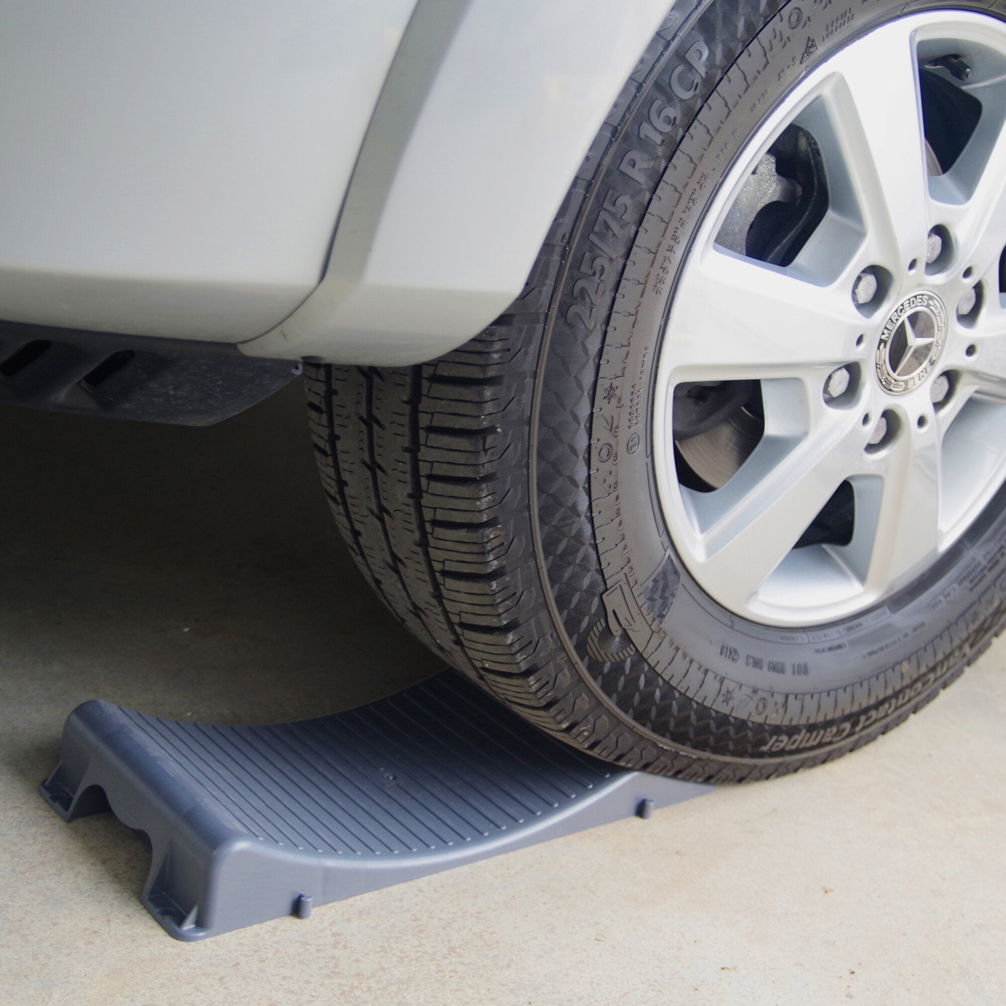 Nahaufnahme eines Autoreifens mit Felge, darunter liegt ein grauer Kunststoff-Unterlegkeil Wheel Saver auf dem Boden
