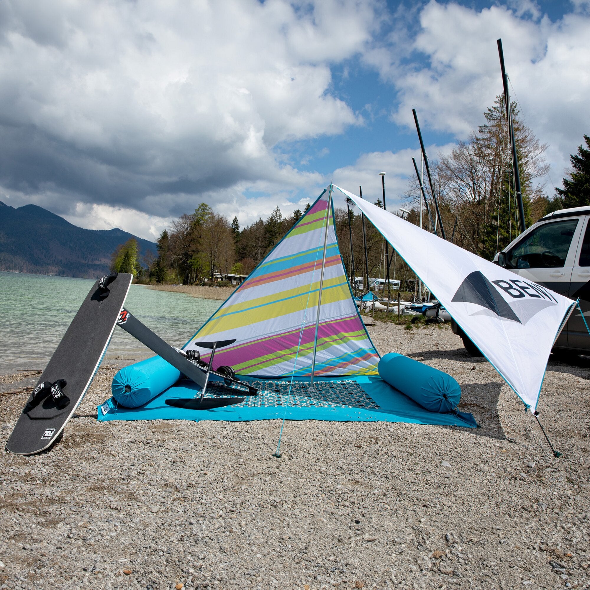 Seeufer mit Kies, bunten Windsurfsegeln und türkisfarbener Zip XL Lounger Kissenrolle sowie aufgeblasenen Polstern im Vordergrund