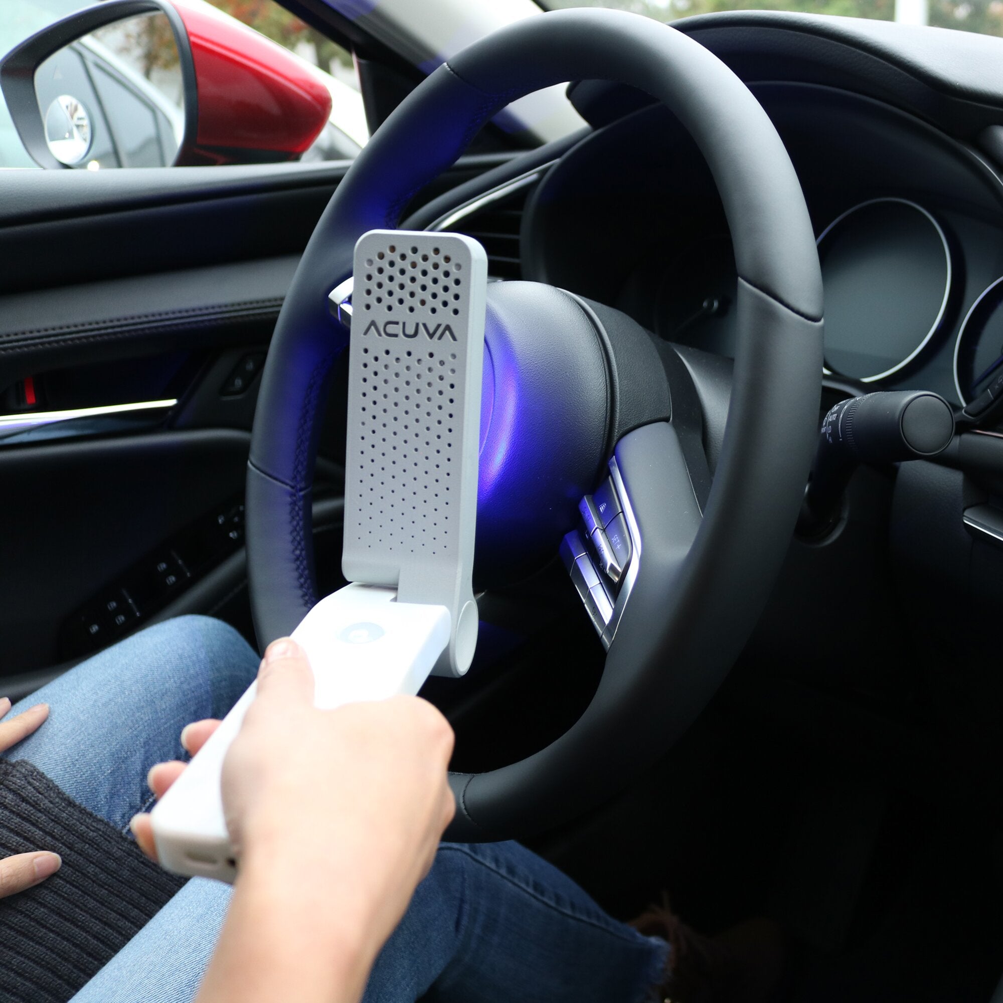 Innenraum eines Autos mit einem grauen, perforierten Desinfektionsgerät von ACUVA am Armaturenbrett neben dem Lenkrad, eine Hand hält einen weißen Gegenstand