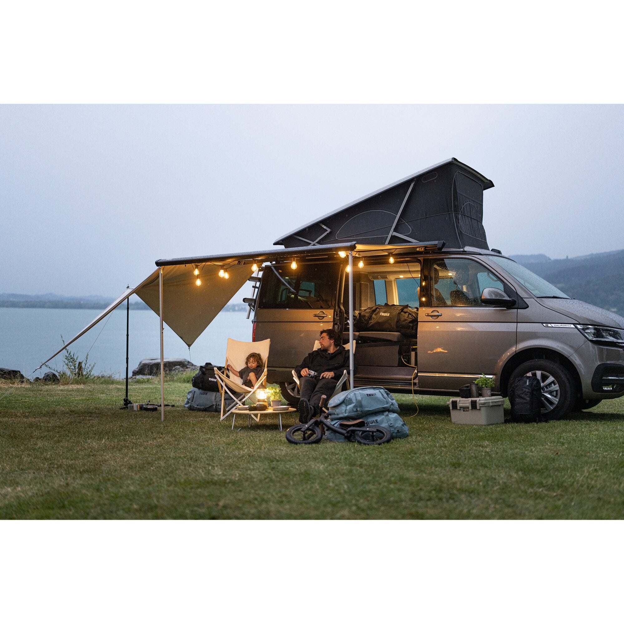 Silberner Campervan mit ausgeklapptem 2 m Seitenvorzelt von Thule Subsola, davor Campingstühle, Tisch und Lichterketten