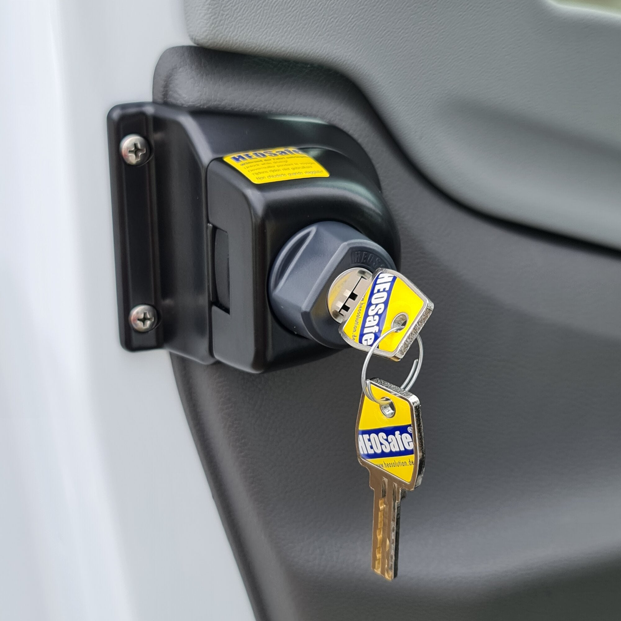 Nahaufnahme eines schwarzen gleichschließenden Zusatzschlosses an einer Türverkleidung, zwei Schlüssel am Schlüsselbund mit gelben HEOSafe Labels