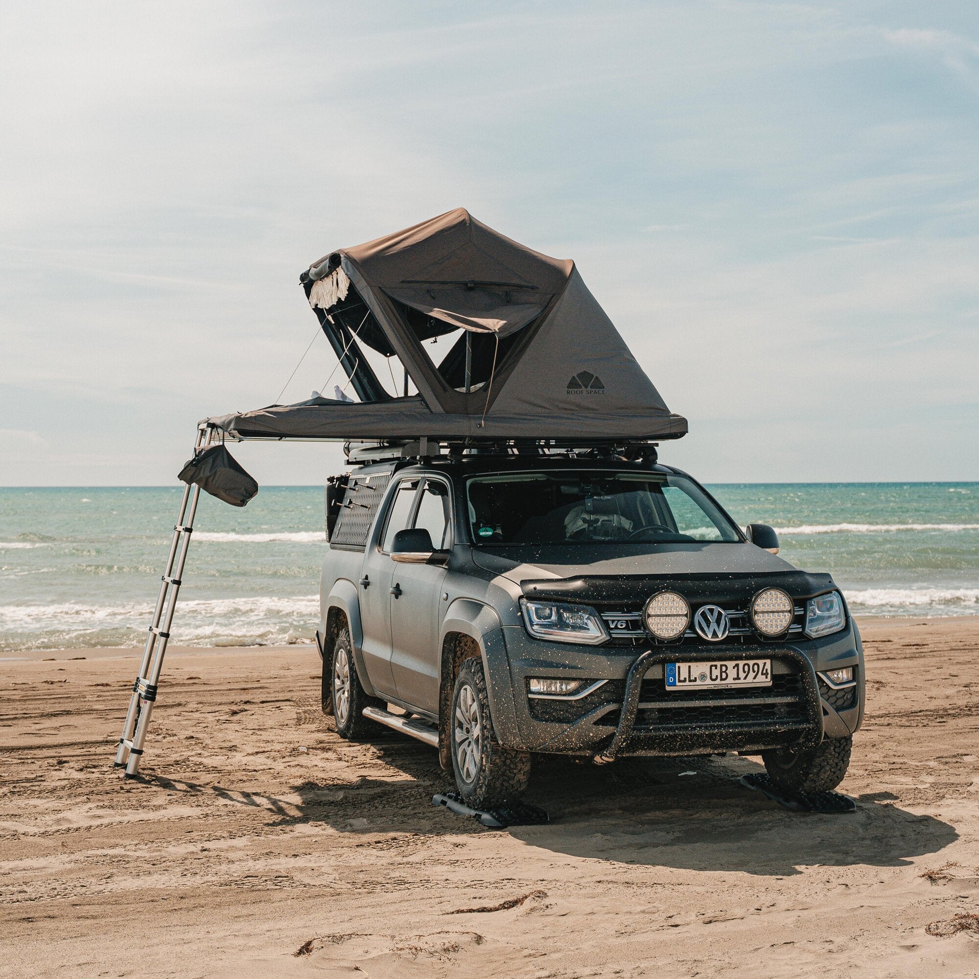 Auf dem Dach eines grauen SUVs steht ein aufgeklapptes Dachzelt Roof Space 2 mit Leiter, am Strand mit Meer im Hintergrund
