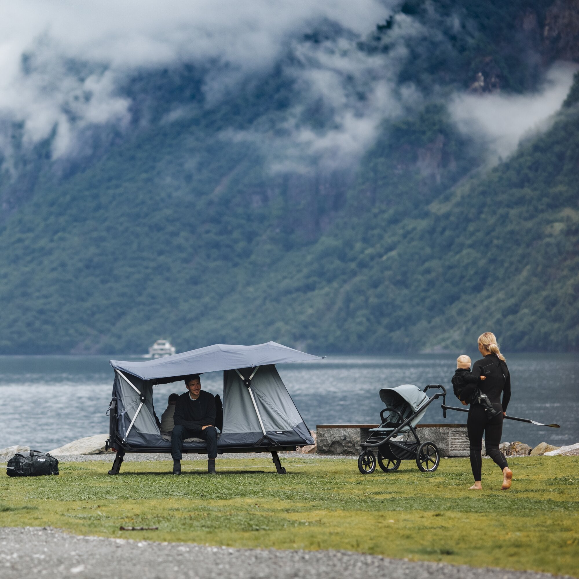 Familie am See neben einem aufgeklappten Thule Outset Anhängerkupplungszelt, graues Zelt auf Gestell, Berglandschaft im Hintergrund