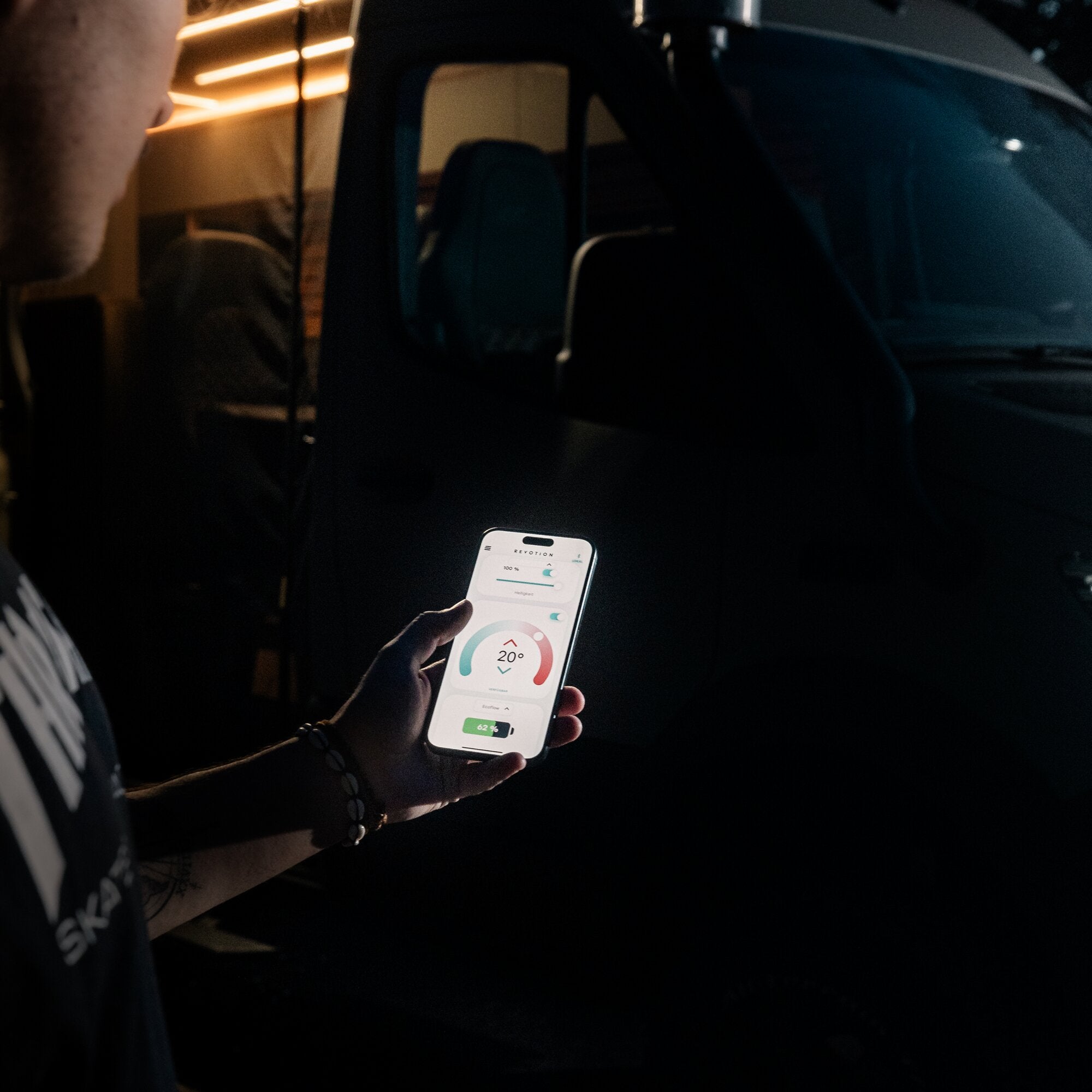 Person hält Smartphone mit kreisförmiger Anzeige (20°) in dunklem Auto-Innenraum; Messgerät liegt auf dem Armaturenbrett, orange Licht