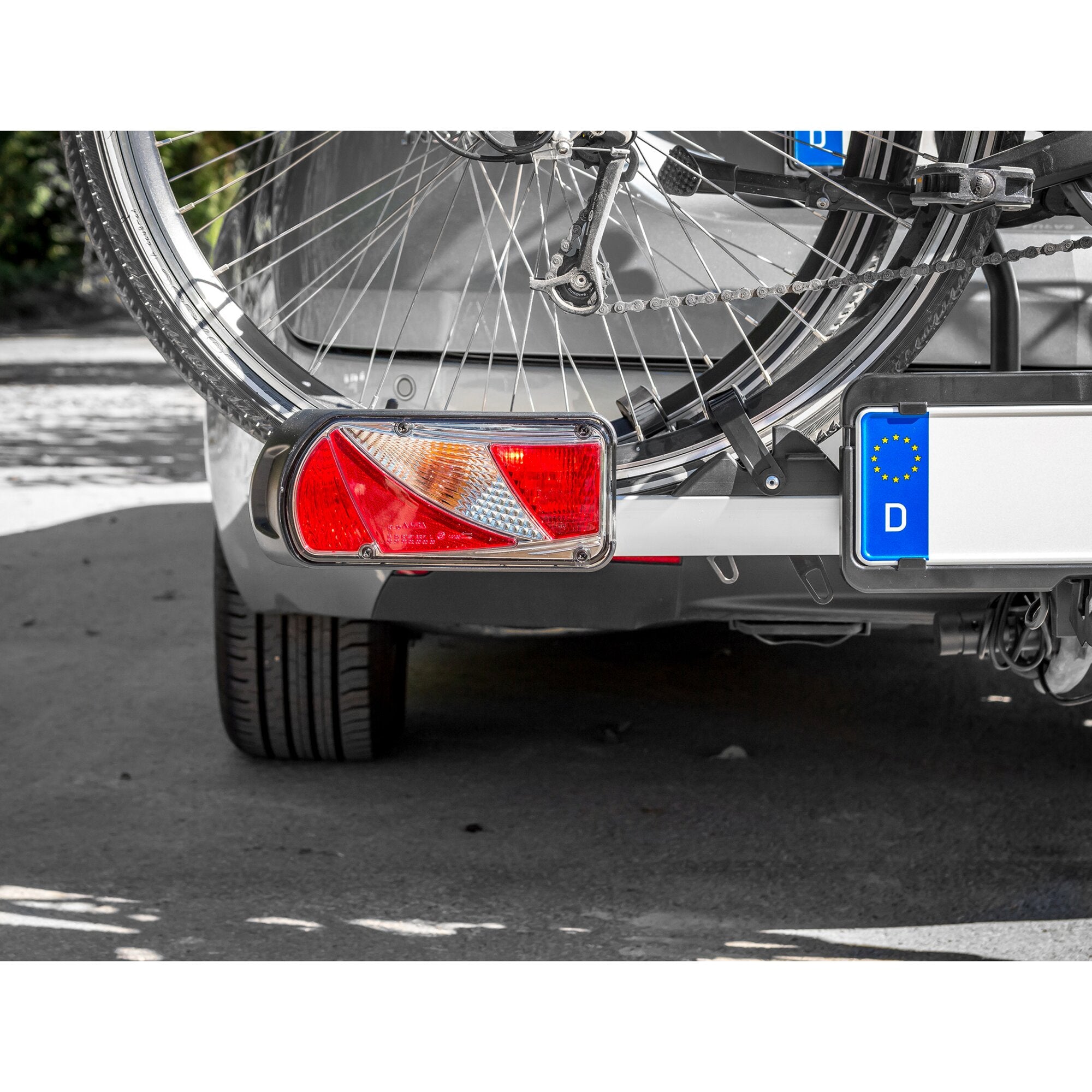 Rückseite eines Autos mit montiertem Fahrradträger an der Anhängerkupplung; sichtbar sind Hinterrad des Fahrrads, Kette, Schaltwerk, Pedal, Heckleuchte und EU-Kennzeichen