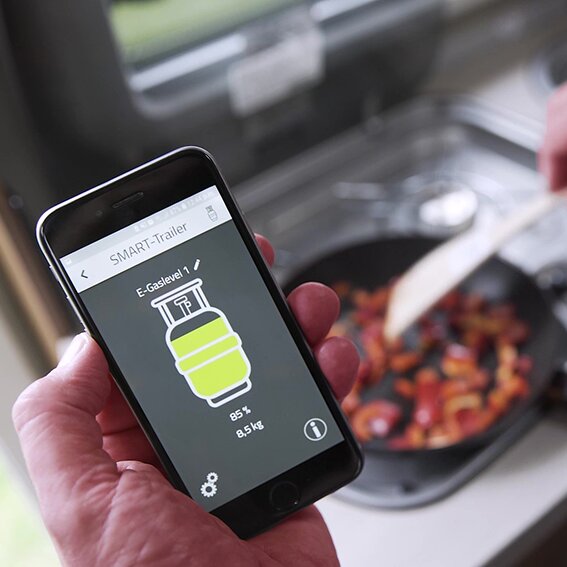 Hand hält Smartphone mit Appanzeige E-Gaslevel; grün leuchtende Gasflasche zeigt 85% und 8.5 kg, Hintergrund Pfanne mit Gemüse
