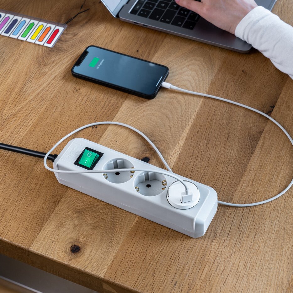 Weiße Steckdosenleiste mit drei runden Steckdosen und grünem Einschaltknopf auf Holztisch; Smartphone wird über USB-Kabel geladen, Laptop im Hintergrund