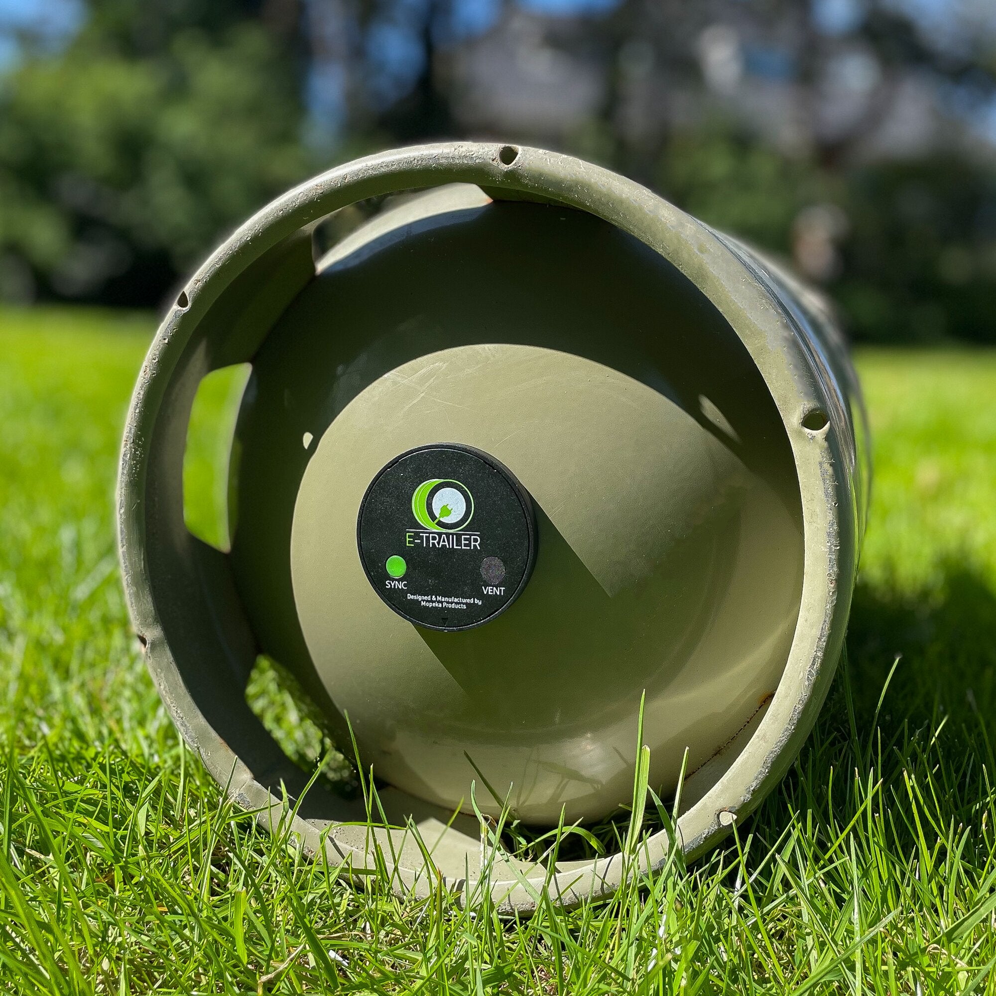 Rundes olivgrünes Gehäuse einer Gas-Füllstandsanzeige mit E-TRAILER Logo, grüner LED-Anzeige und SYNC/VENT-Hinweisen, Gras im Hintergrund
