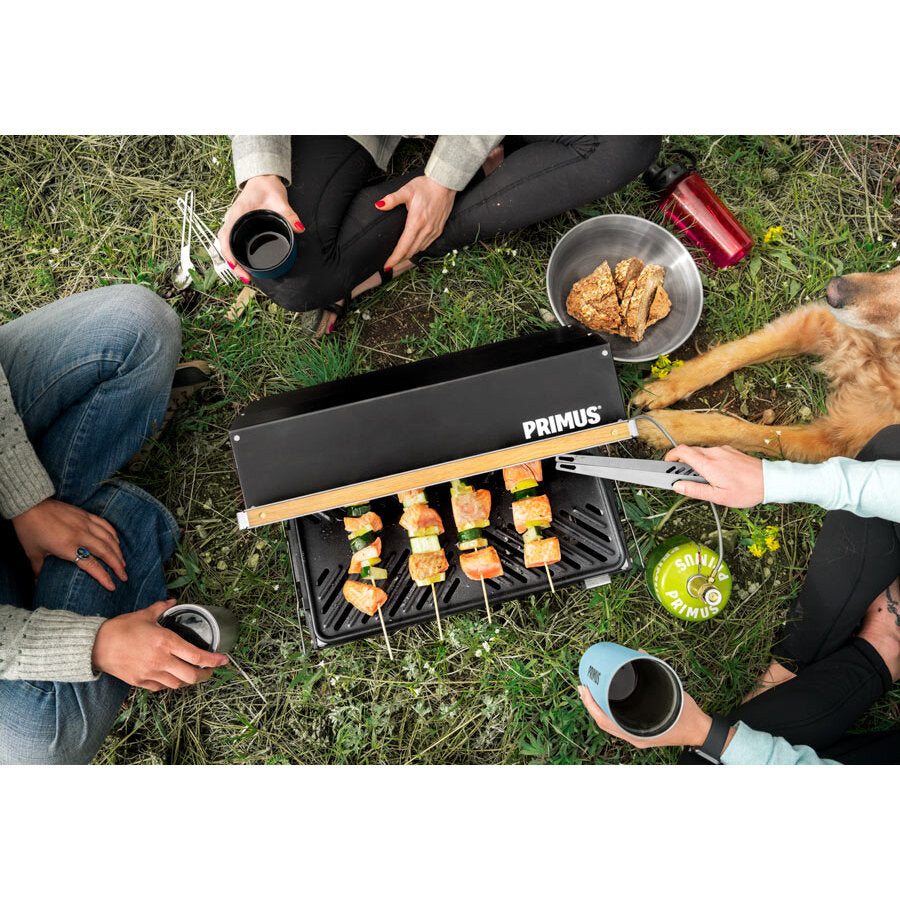 Gasgrill Kuchoma 2500 Watt offen auf Gras; Spieße mit Fleisch und Gemüse grillen, Primus-Deckel, Gasflasche daneben, Leute sitzen, Hund dabei