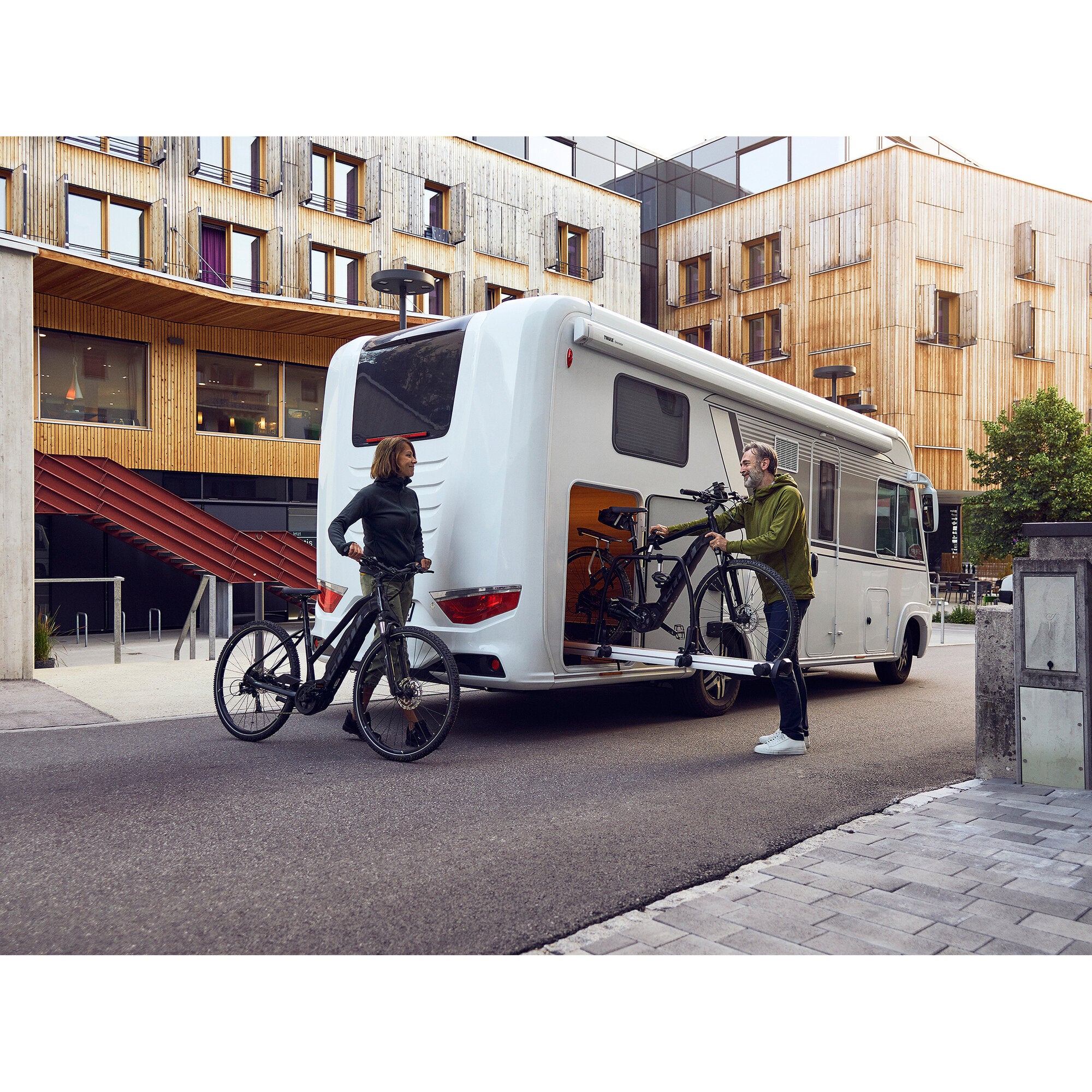 Weißes Wohnmobil mit Thule VeloSlide Fahrradträger am Heck, zwei Fahrräder gesichert, zwei Personen davor vor moderner Holzarchitektur