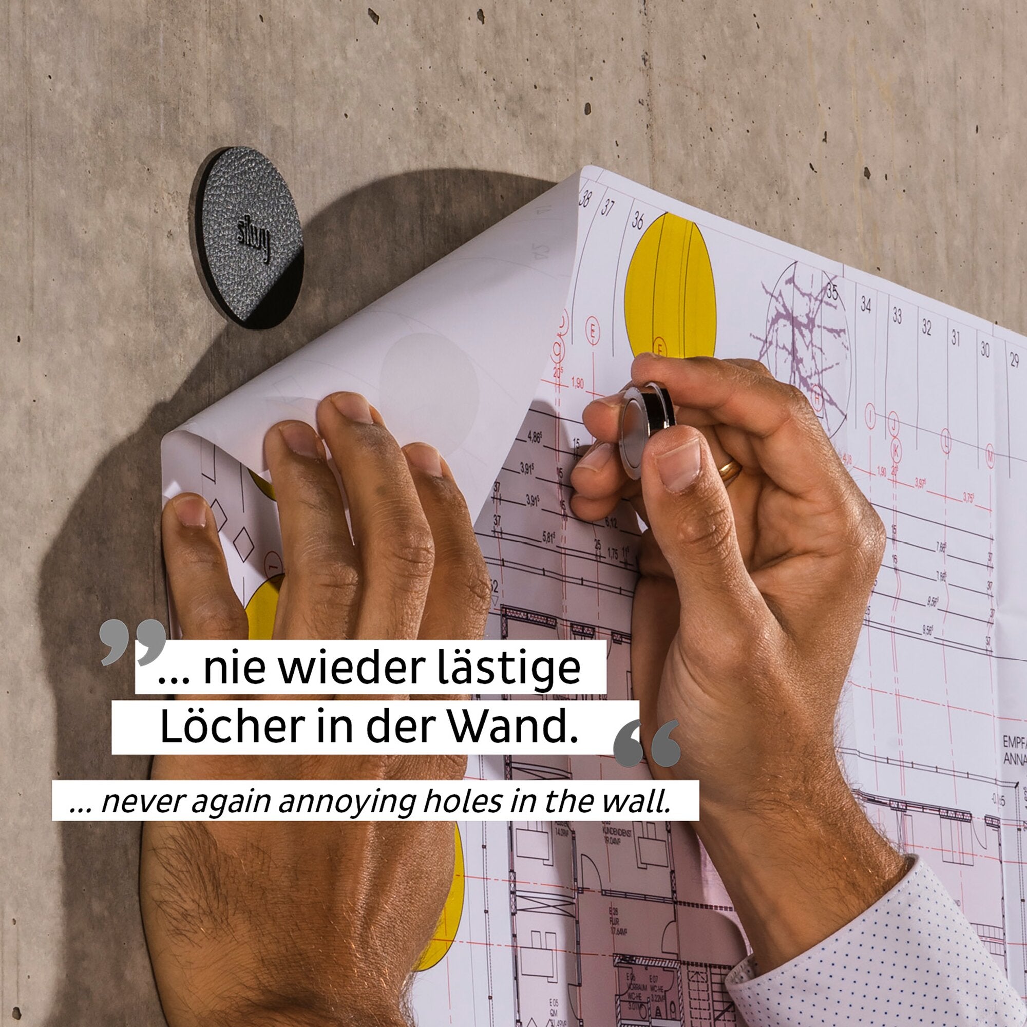 Runder Magnet-Pins SMART aus Metall mit strukturierter Oberfläche und dunklem Rand liegt auf Beton, Hände halten ein Blatt mit Zeichnungen