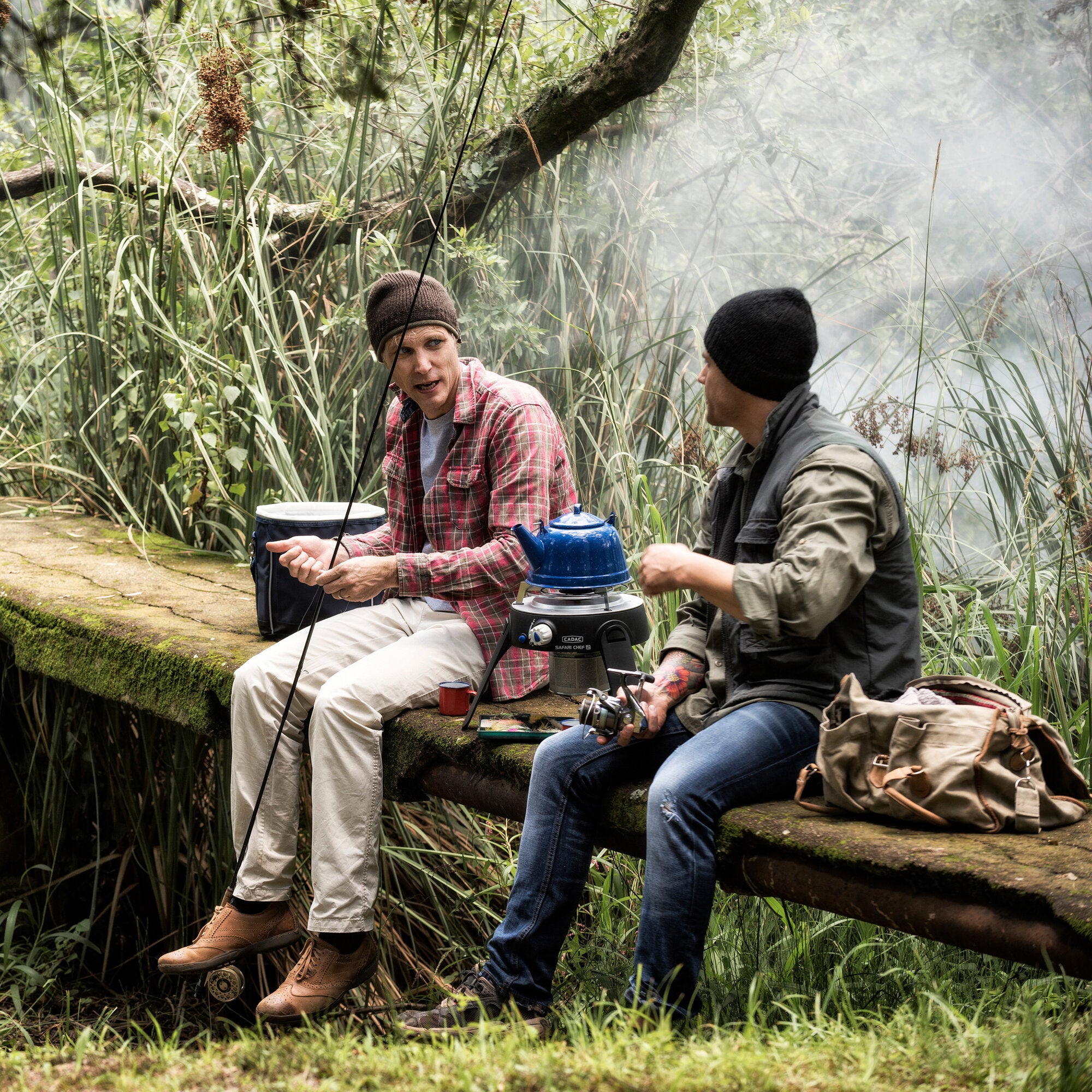 Zwei Männer im Freien sitzen an einer moosigen Steinbank und nutzen einen Cadac Safari Chef 2 Gasgrill mit blauem Topf; rote Tasse vorne