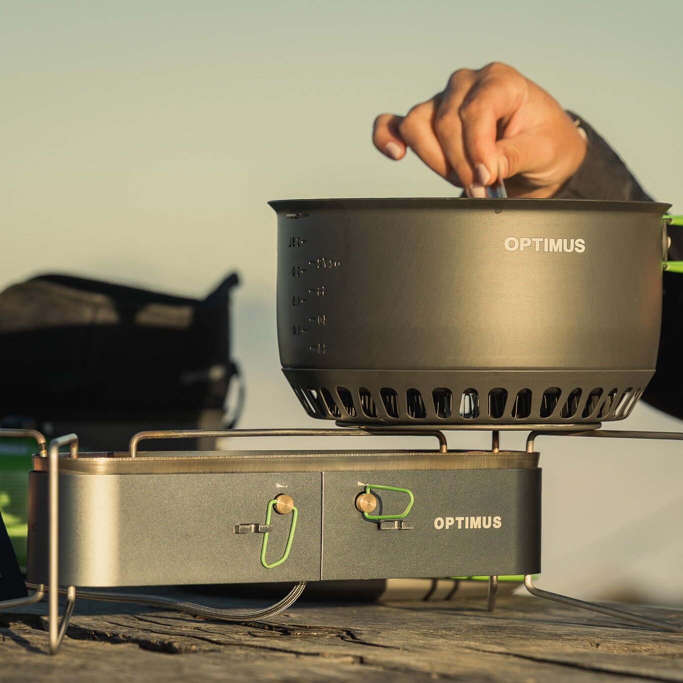 Gaskocher Gemini II mit Topf, Hand greift Tragegriff am Topf, sichtbar OPTIMUS-Logo auf Topf und Basis, Outdoor-Kochset