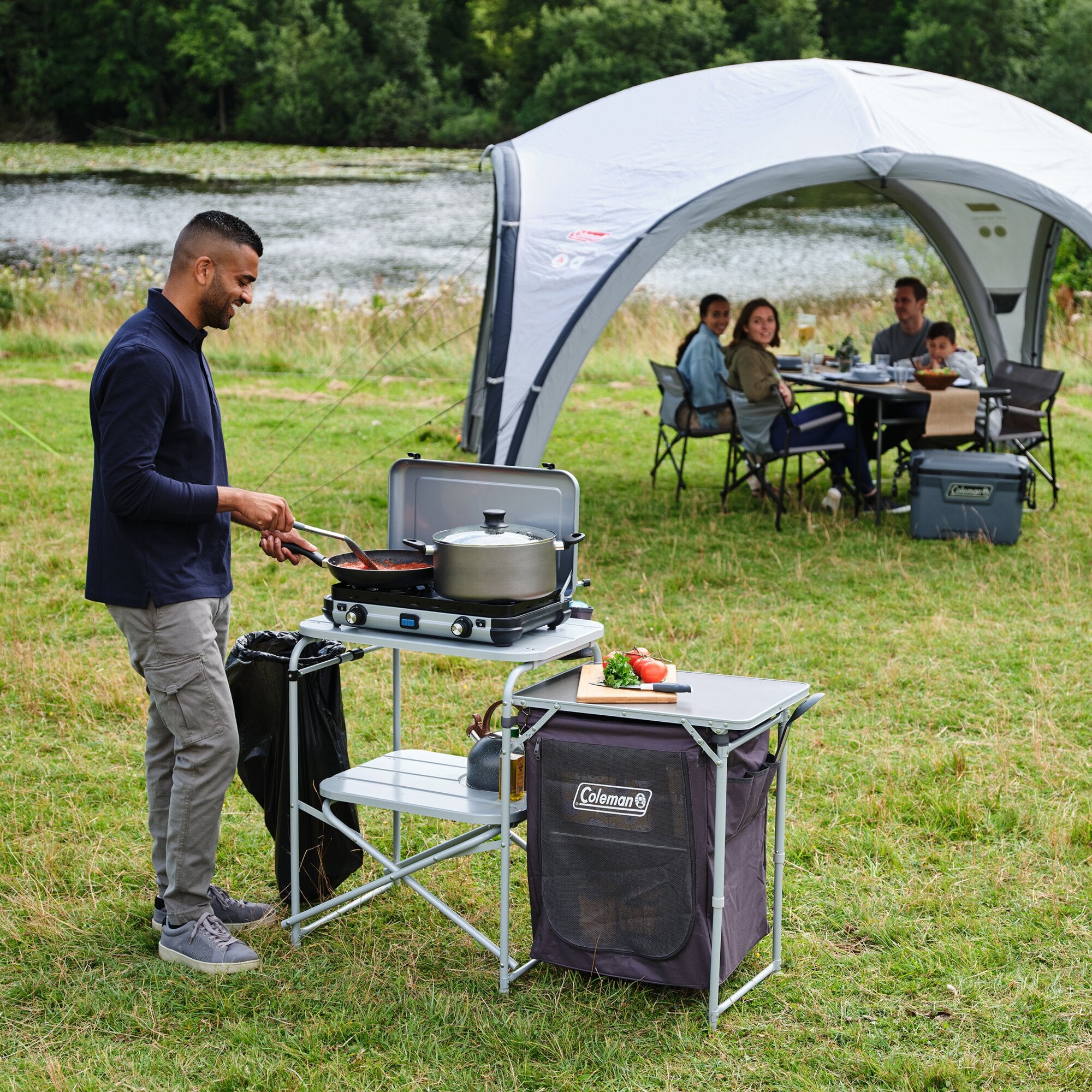 Mann kocht auf einer grauen Campingküche Cooking Stand mit zwei Brennern; davor Schneidebrett mit Gemüse, Gruppe sitzt unter einem Pavillon