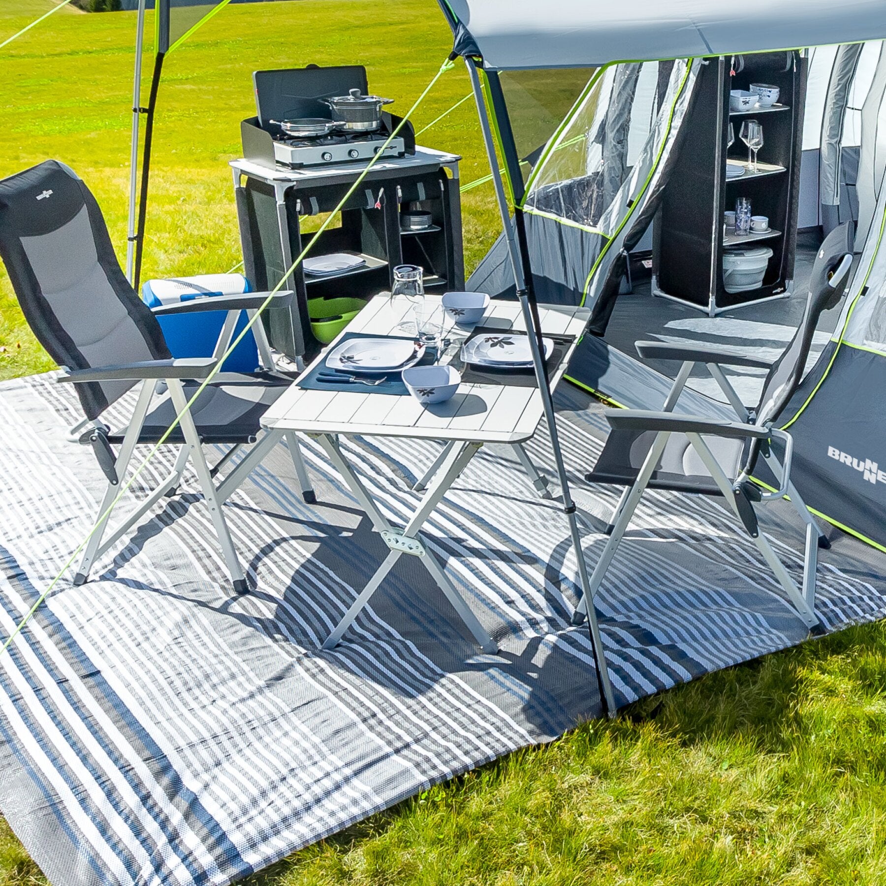 Rolltisch Silver Gapless 140 cm als Camping-Essbereich mit zwei Klappstühlen, Zweibrenner-Grill, Geschirrregal und gestreiftem Teppich auf Gras