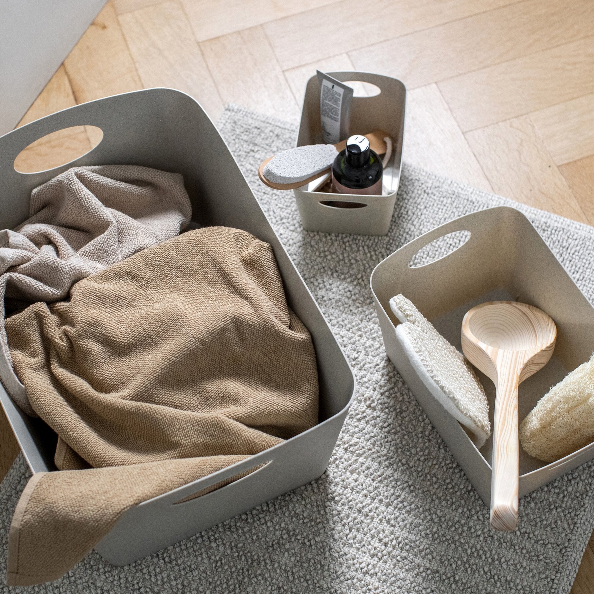 Sandfarbene Aufbewahrungsbox BOXXX M mit Griffen auf hellgrauem Teppich; innendrin beige Stoffe und eine Pumpspenderflasche sichtbar