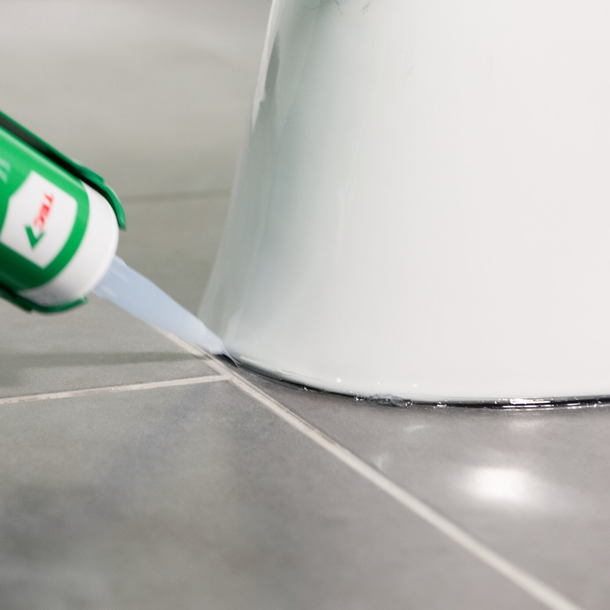 Nahaufnahme einer TRANSCLEAR Kleb- und Dichtstoff-Kartusche mit grüner Kappe, an einer glatten weißen Oberfläche, neben einer Silikonnaht am Boden