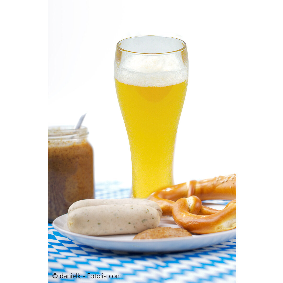 Langes Weißbierglas mit goldgelbem Bier und Schaum, davor Teller mit Weißwürsten, Laugenbrezel und Aufstrich, blau-weiß kariertes Tischtuch im Hintergrund