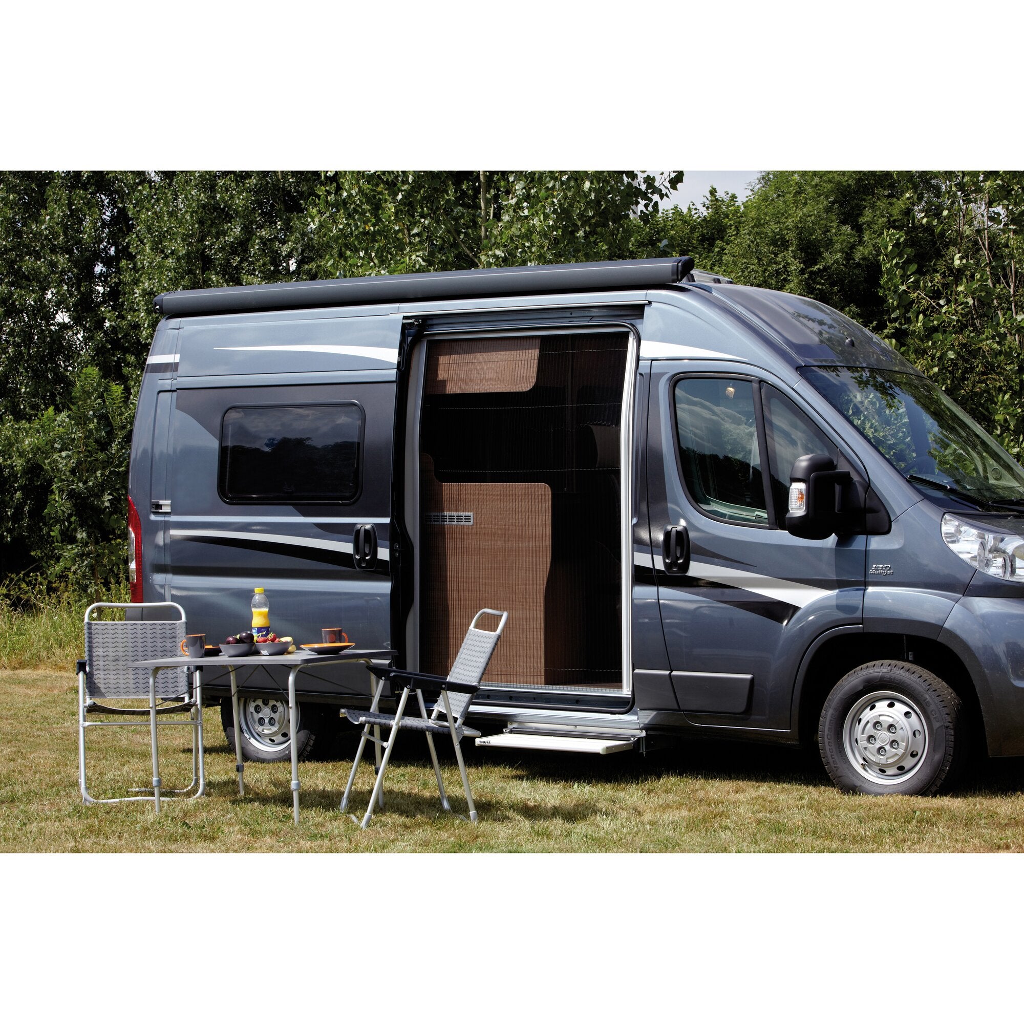 Graues Wohnmobil mit offener Seitentür; auf dem Dach ein langer schwarzer Insektenschutz FlyTec FT 200; davor Campingtisch mit Stühlen und Obst, grüner Hintergrund