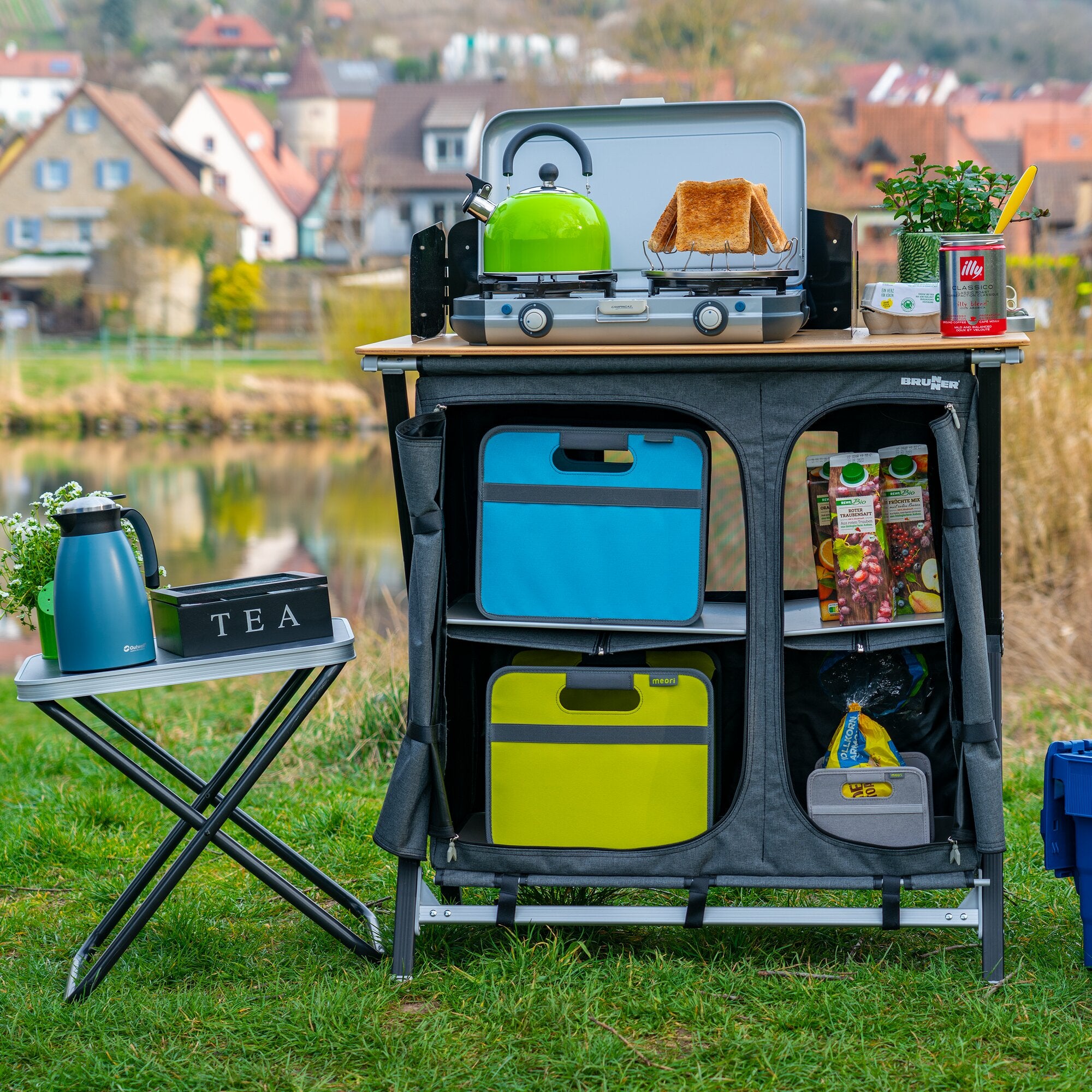 Offenes graues Cross Cooker HWT SQ Zwei-Brenner Outdoor-Kochsystem auf Tisch, grüner Kessel links, Toastbrot auf dem Rost rechts, Hintergrund Gartenlandschaft